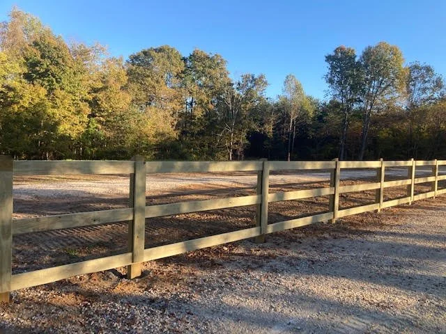 A wooden fence along a gravel path with trees in the background under a clear blue sky.
