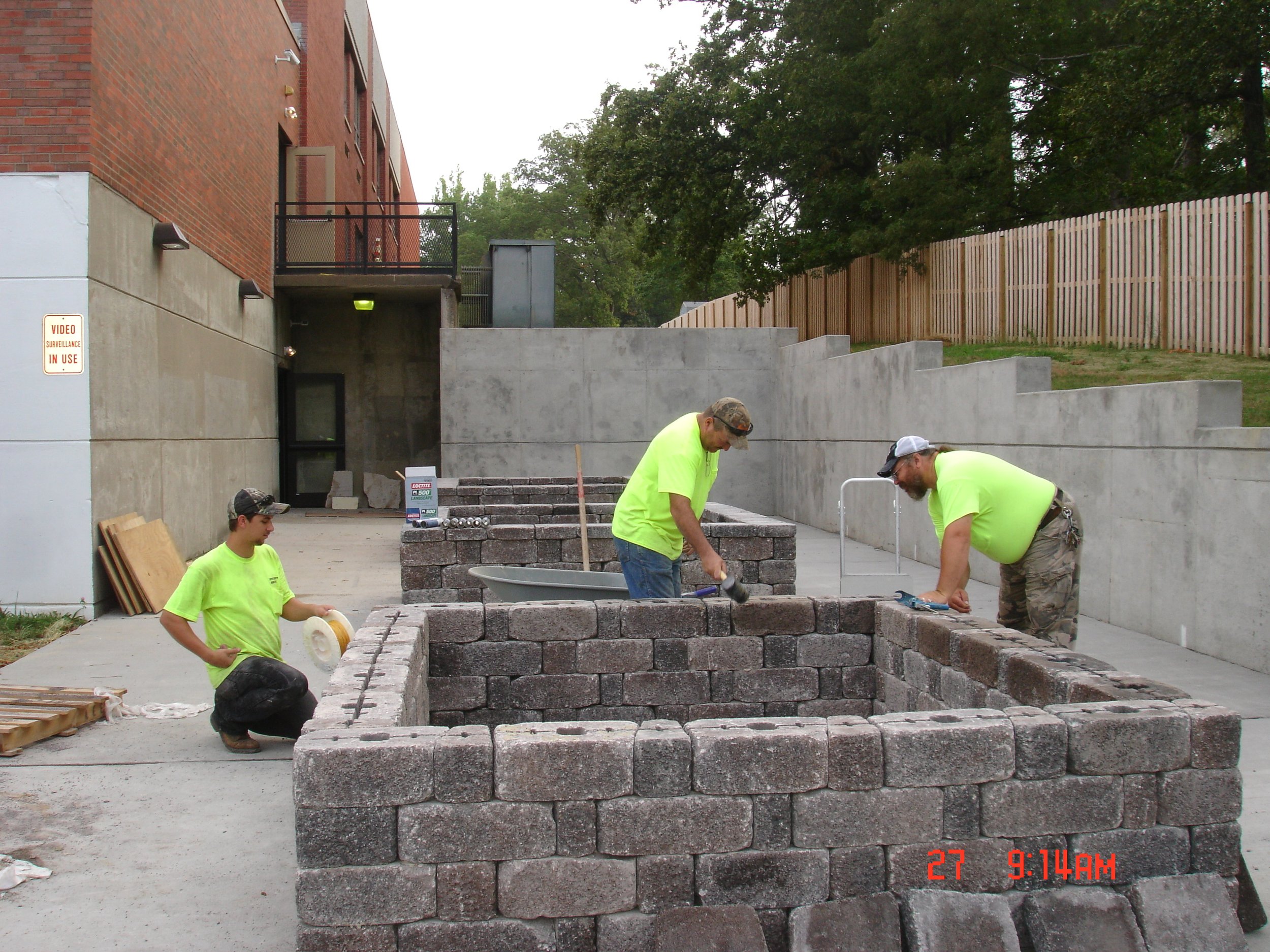 Three construction workers building a brick structure outdoors on a paved area next to a brick building and a wooden fence, with trees in the background.