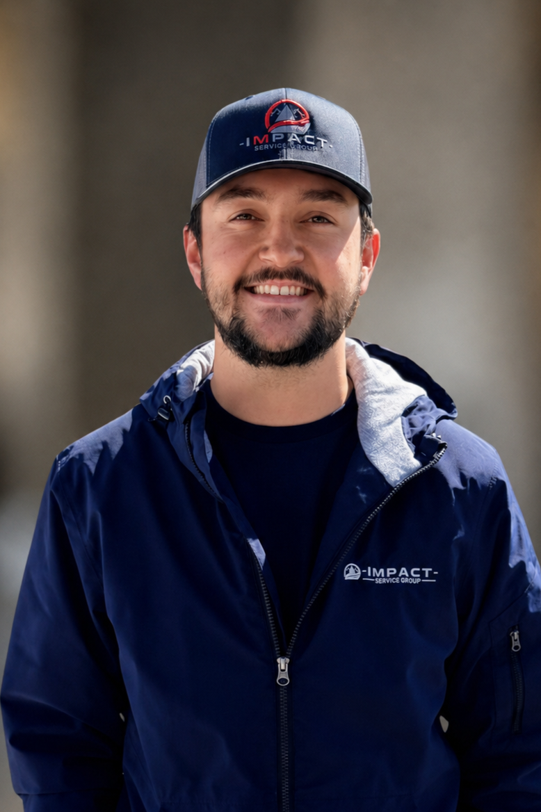 A man smiling outdoors, wearing a navy blue polo shirt and a matching cap, both with a logo that says "Impact Heating & Air."