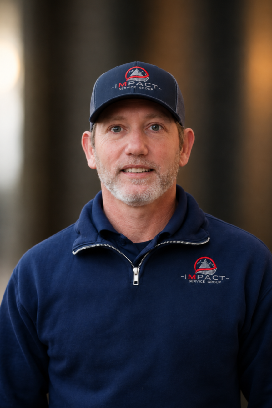 A man wearing a navy blue jacket and matching cap with 'Impact Service Group' logo standing on a city sidewalk near brick buildings.