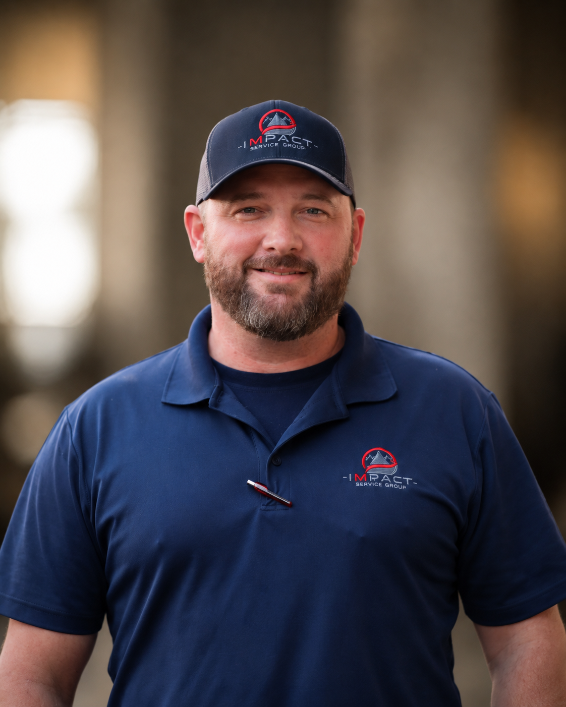 A man with a beard and short hair wearing a navy blue polo shirt and a matching cap, both with the 'Impact Service Group' logo, standing outside on a sidewalk with a brick building and windows in the background.