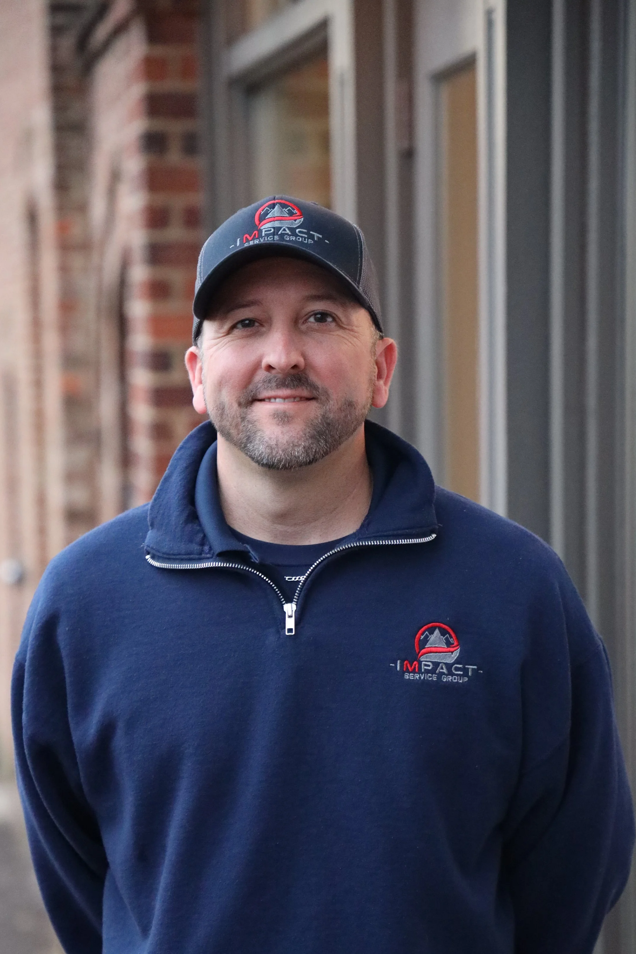 Man wearing a navy blue Impakt Service Group sweatshirt and cap, standing outdoors near a building with brick and metal siding.