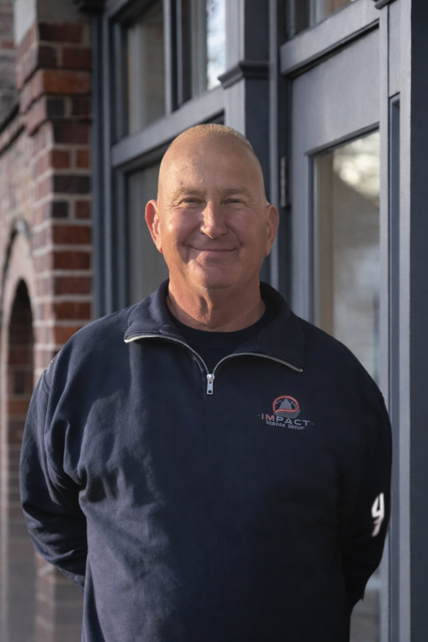 A smiling man stands outside in front of a gray building with glass windows, wearing a black zip-up jacket with a logo that says 'IMPACT' on the chest.