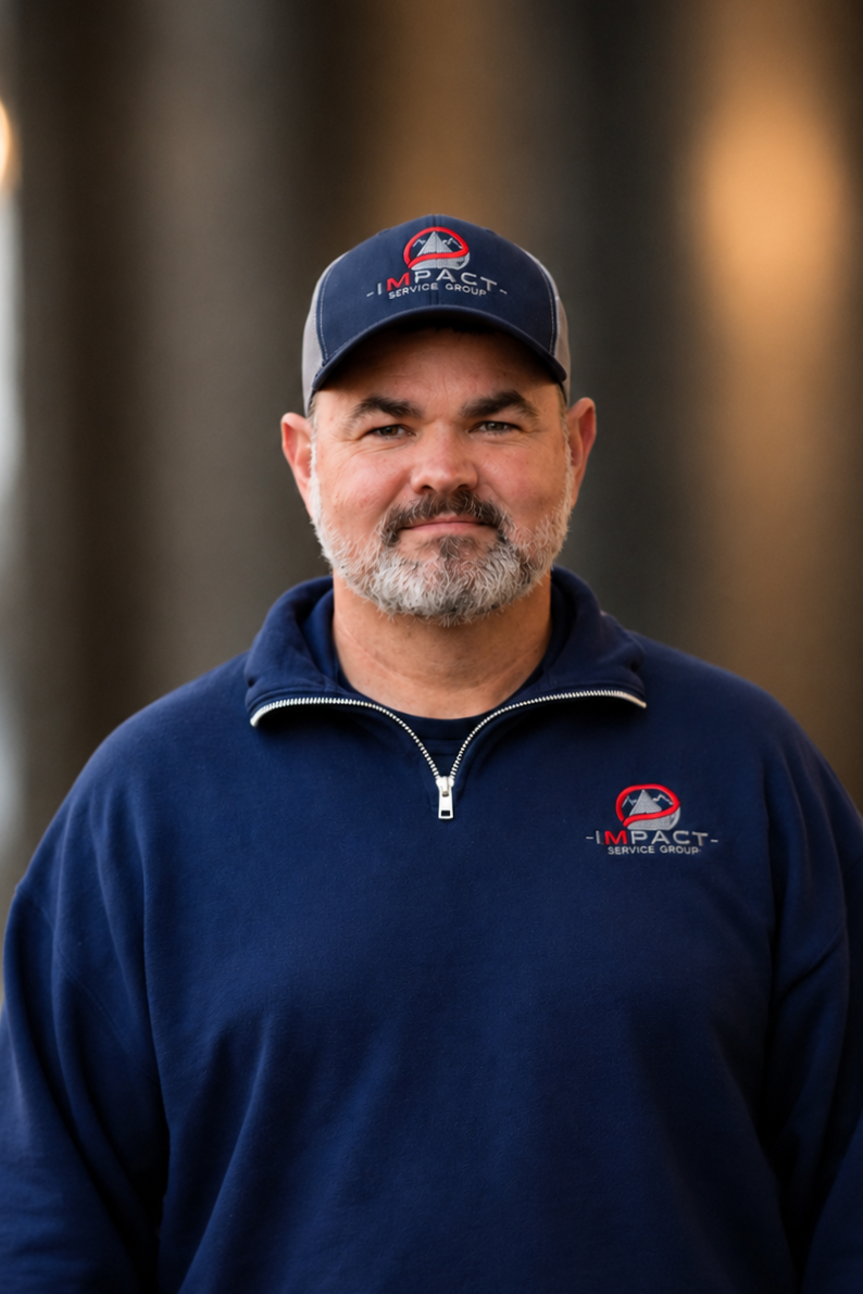 A man with a beard wearing a dark blue sweatshirt and a matching cap with the 'Impact Service Group' logo, standing outdoors in front of a brick wall and a building with large windows.