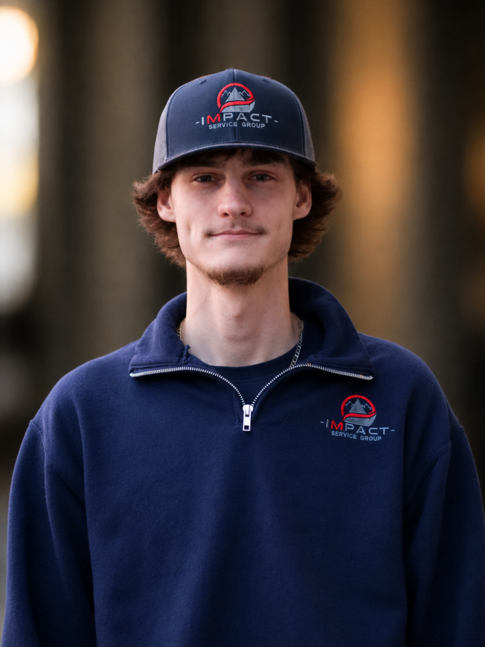 A young man wearing a dark blue baseball cap and matching navy fleece jacket with the 'Impact Service Group' logo on both the cap and jacket, standing outside near a brick building with windows.