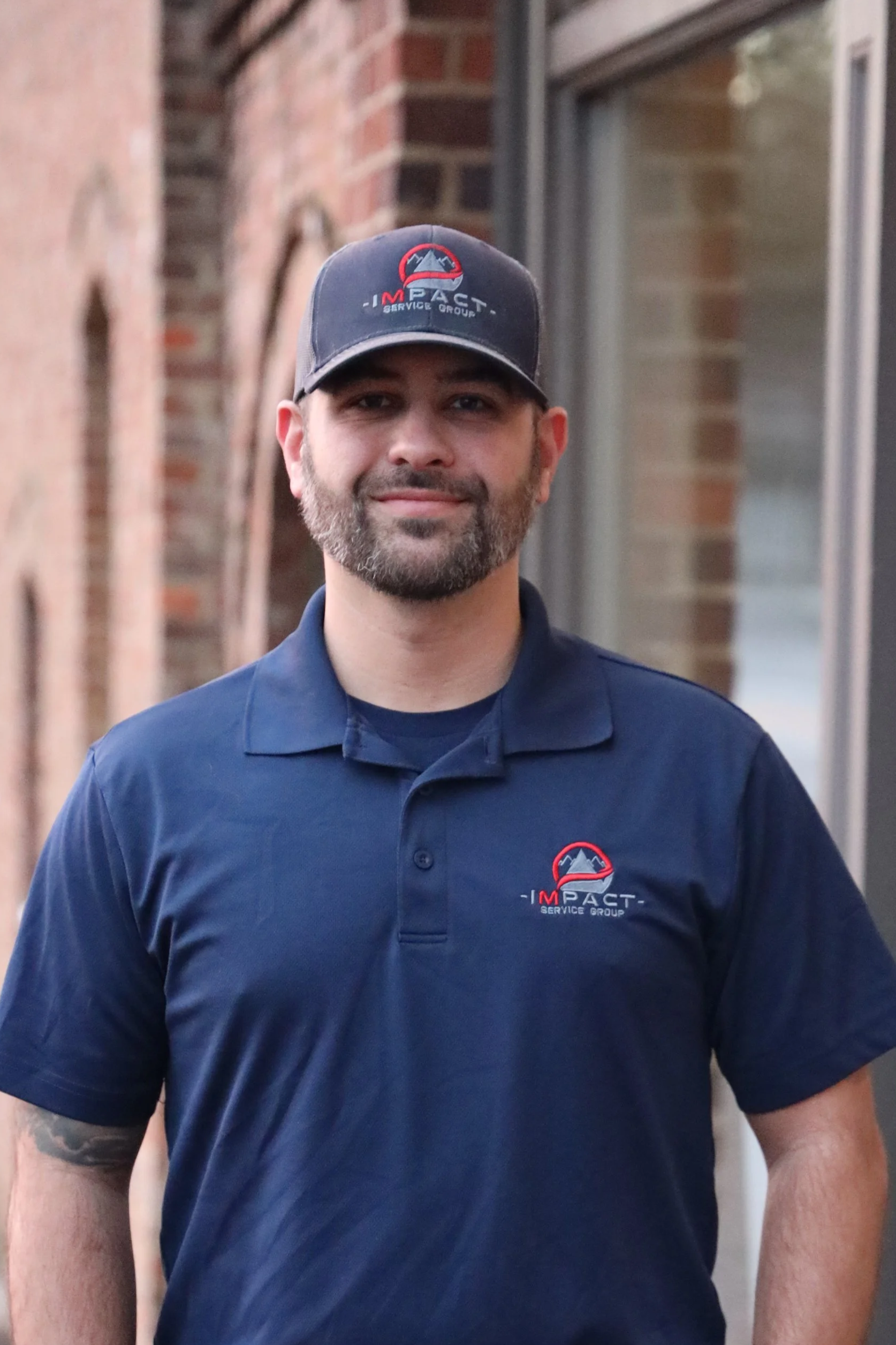 A man with a beard and mustache wearing a navy blue polo shirt and matching cap, both with a logo for 'Impact Service Group,' standing outdoors in front of brick walls.