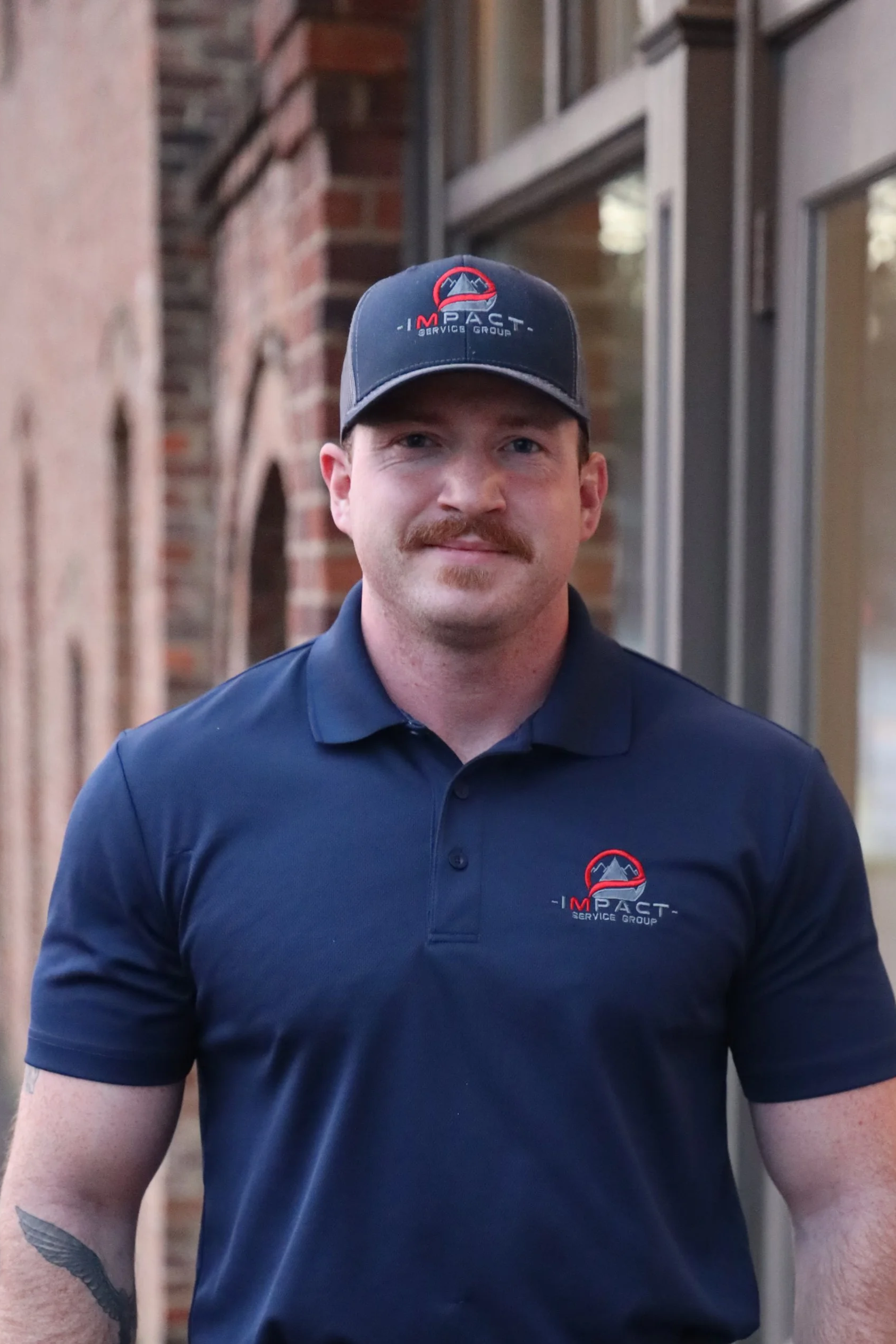 A man with a mustache and short hair wearing a navy polo shirt and a cap, both with the ImPACT Service Group logo, standing outside with brick buildings in the background.