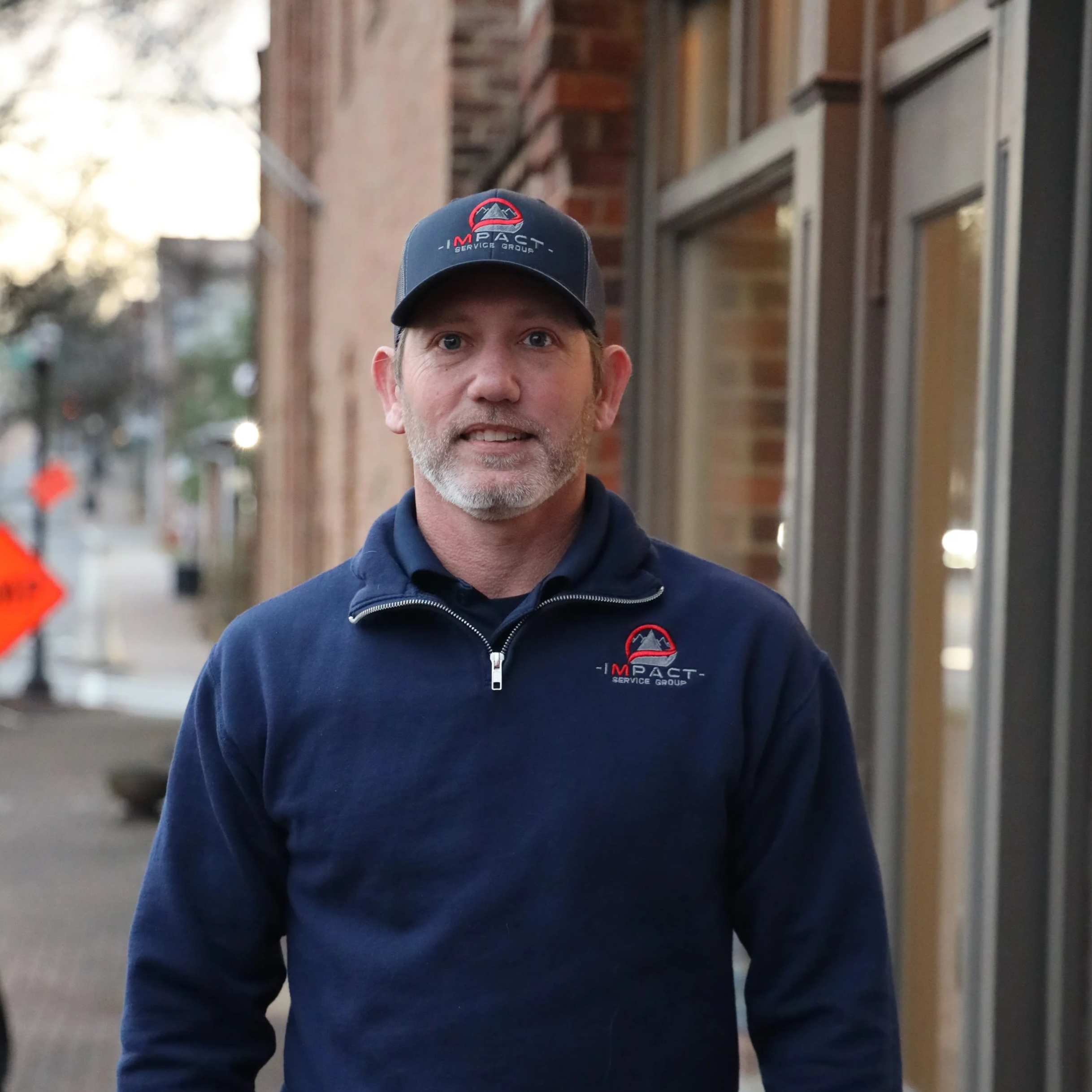 A man wearing a navy blue jacket and matching cap with 'Impact Service Group' logo standing on a city sidewalk near brick buildings.