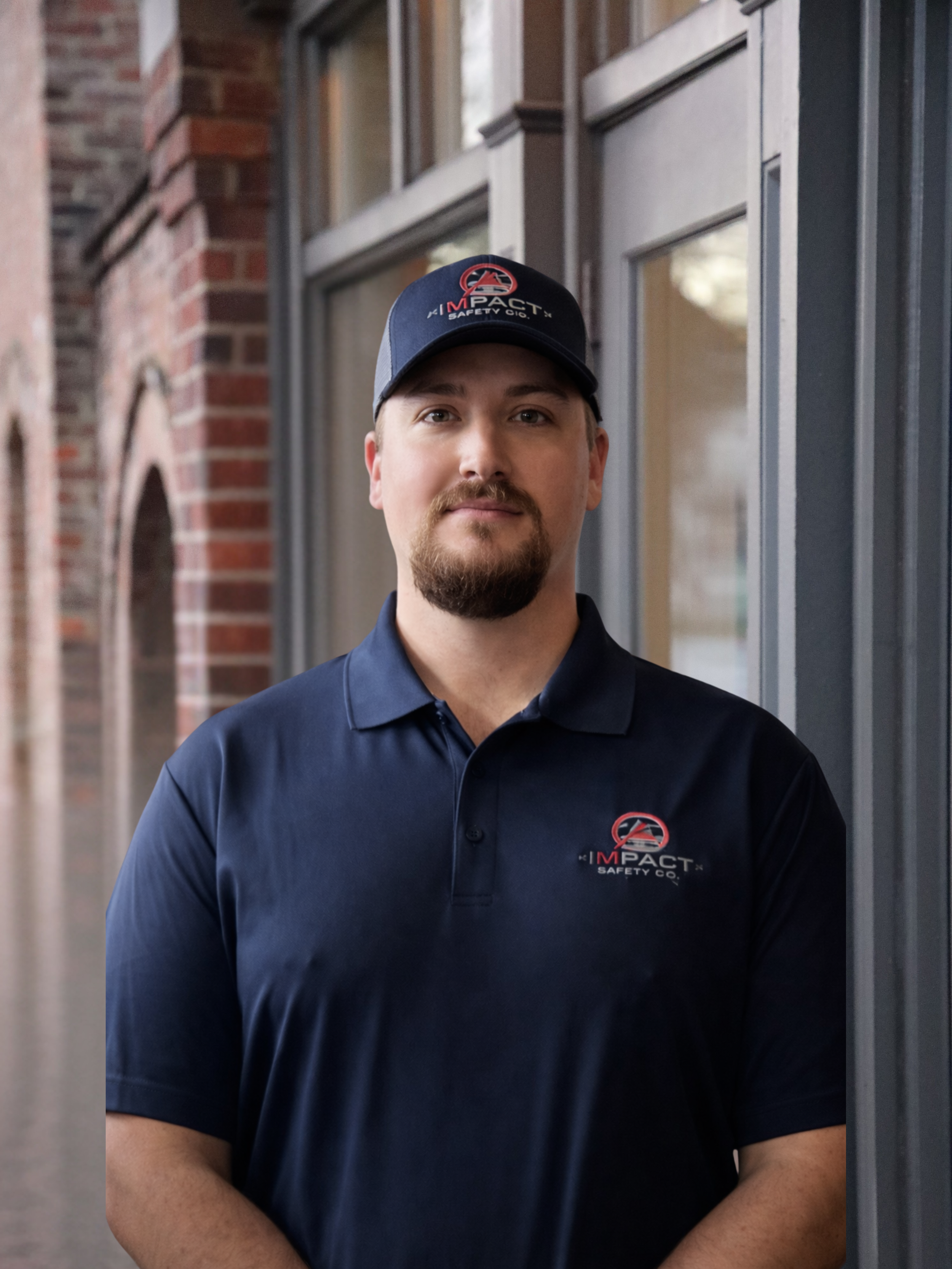 A man in a navy blue polo shirt and matching cap with a logo from Impact Safety Co. standing outside in front of a brick building with large windows.