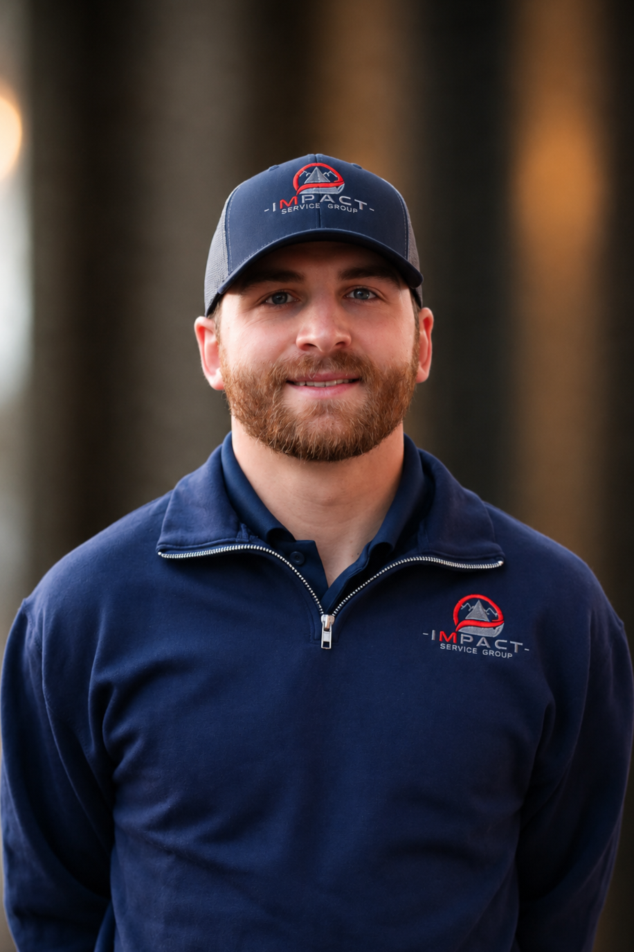 A man with a beard wearing a navy blue jacket and a cap with an 'Impact Service Group' logo, standing outdoors near a brick building.