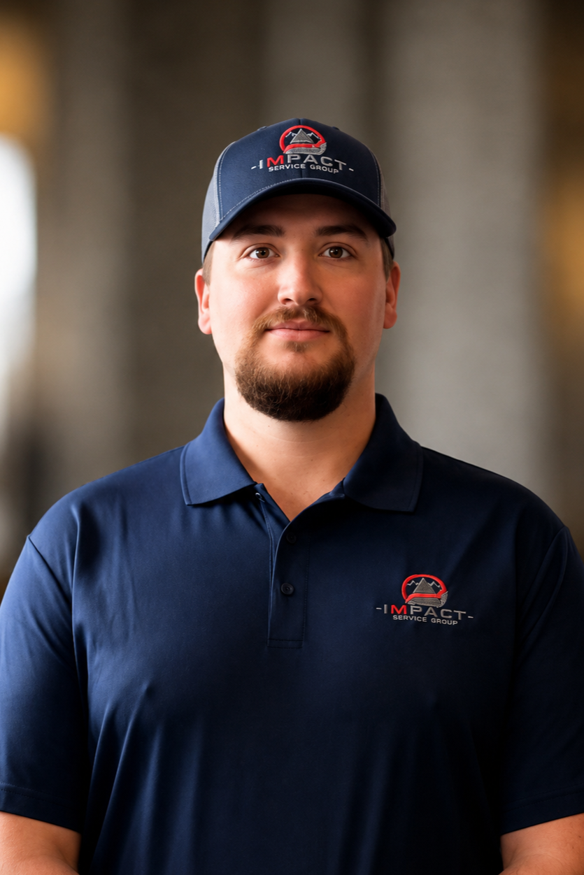 A man in a navy blue polo shirt and matching cap with a logo from Impact Safety Co. standing outside in front of a brick building with large windows.
