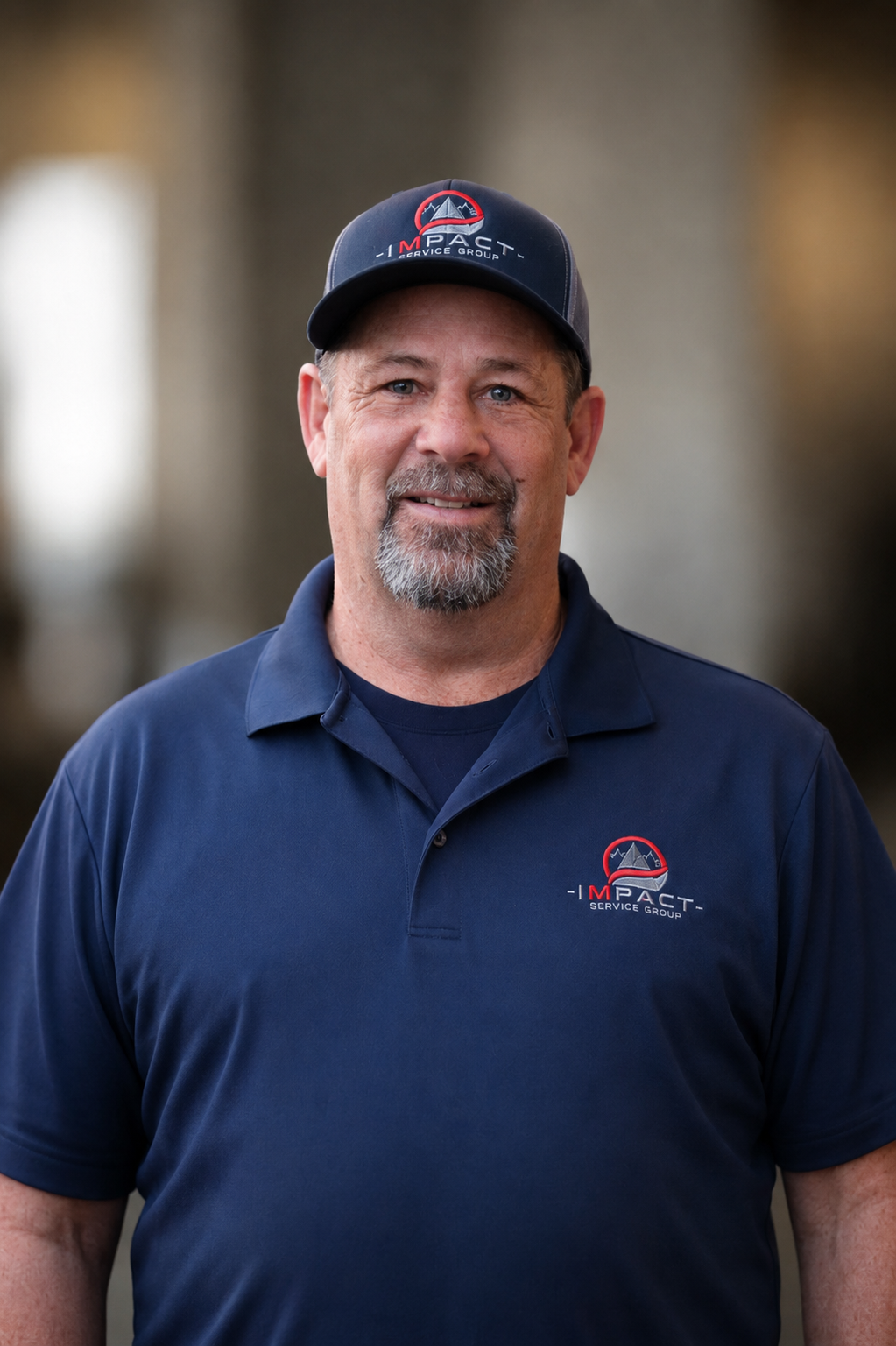 A man with a beard and mustache wearing a dark cap and navy polo shirt, both with the 'Impact Service Group' logo, standing outdoors near a brick building with large windows.