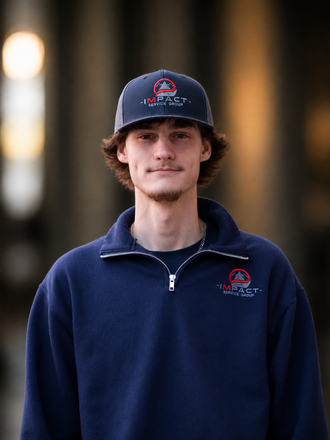 A young man wearing a dark blue baseball cap and matching navy fleece jacket with the 'Impact Service Group' logo on both the cap and jacket, standing outside near a brick building with windows.