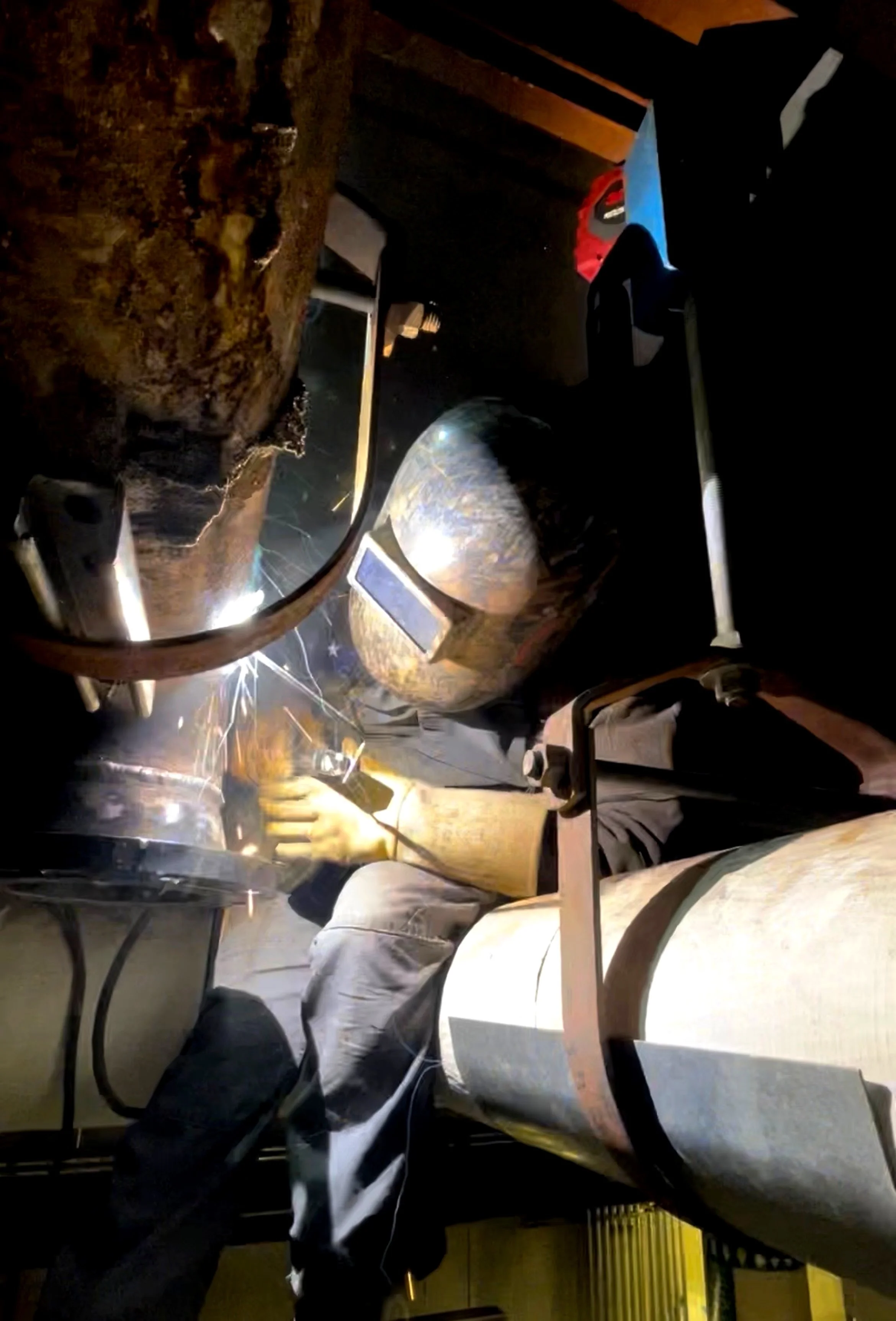 Photo taken inside a workshop showing a person welding metal with sparks flying, wearing protective gear including a welding helmet and gloves, with various tools and materials around.