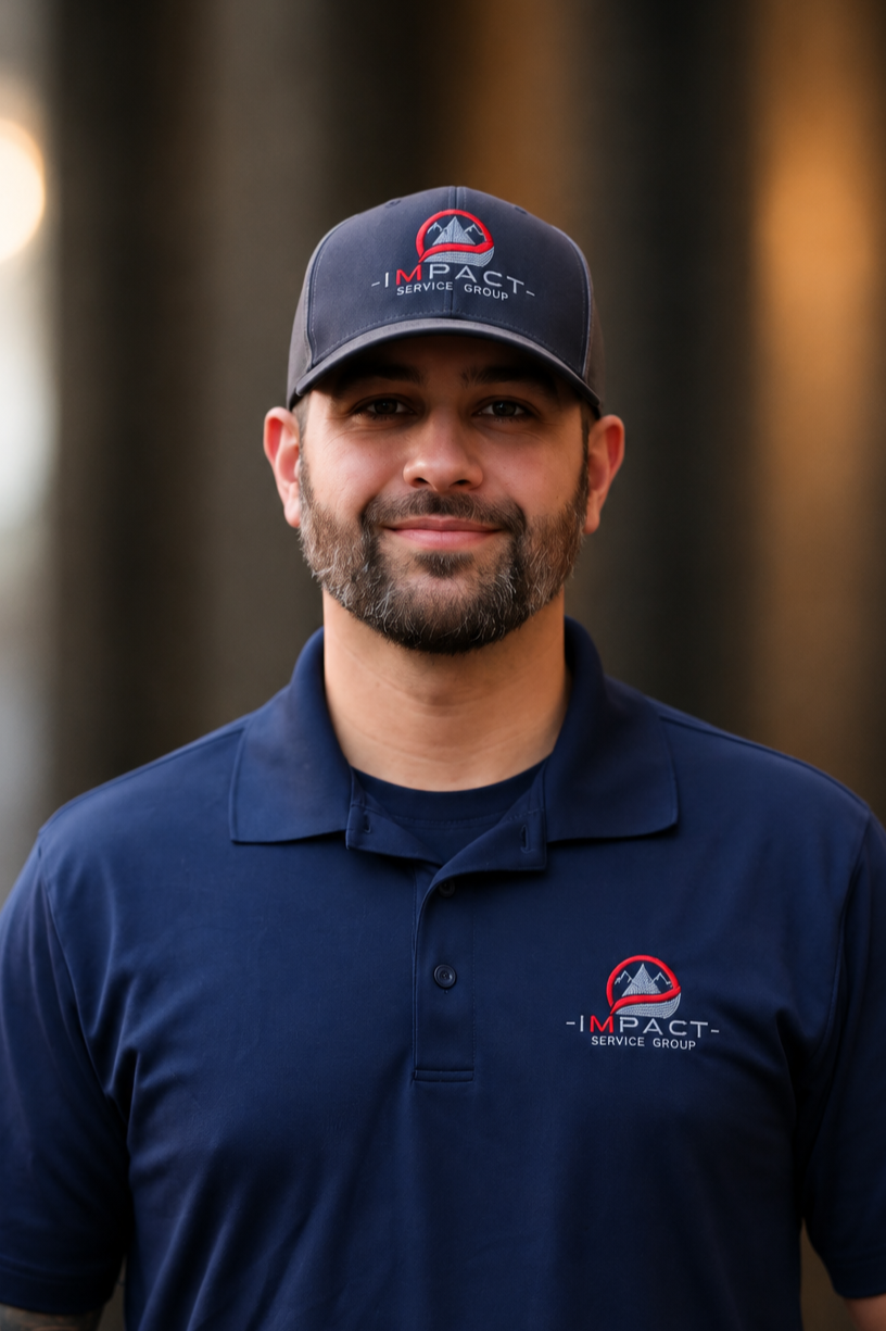 A man with a beard and mustache wearing a navy blue polo shirt and matching cap, both with a logo for 'Impact Service Group,' standing outdoors in front of brick walls.