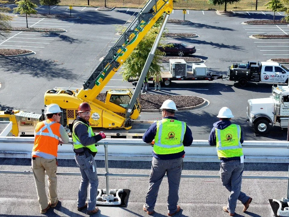 Construction workers and engineers in safety vests and helmets standing on a platform observing construction work on a bridge or overpass. A yellow crane and various construction vehicles are visible below, with a parking lot in the background.