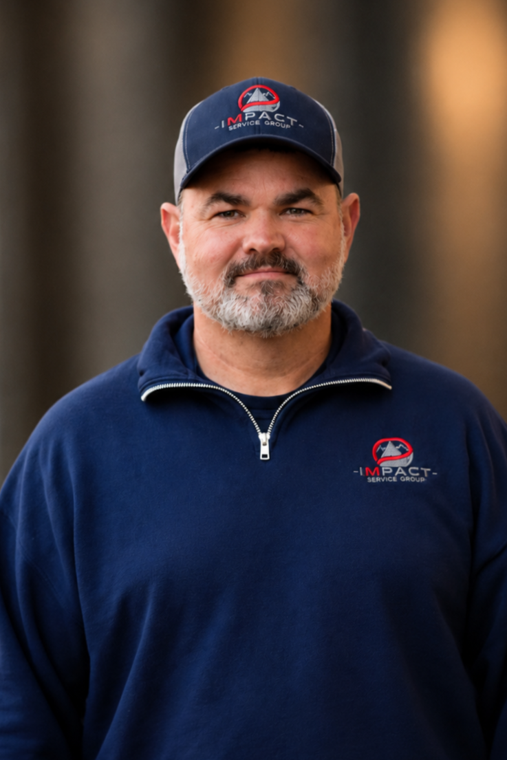 A man with a beard wearing a dark blue sweatshirt and a matching cap with the 'Impact Service Group' logo, standing outdoors in front of a brick wall and a building with large windows.