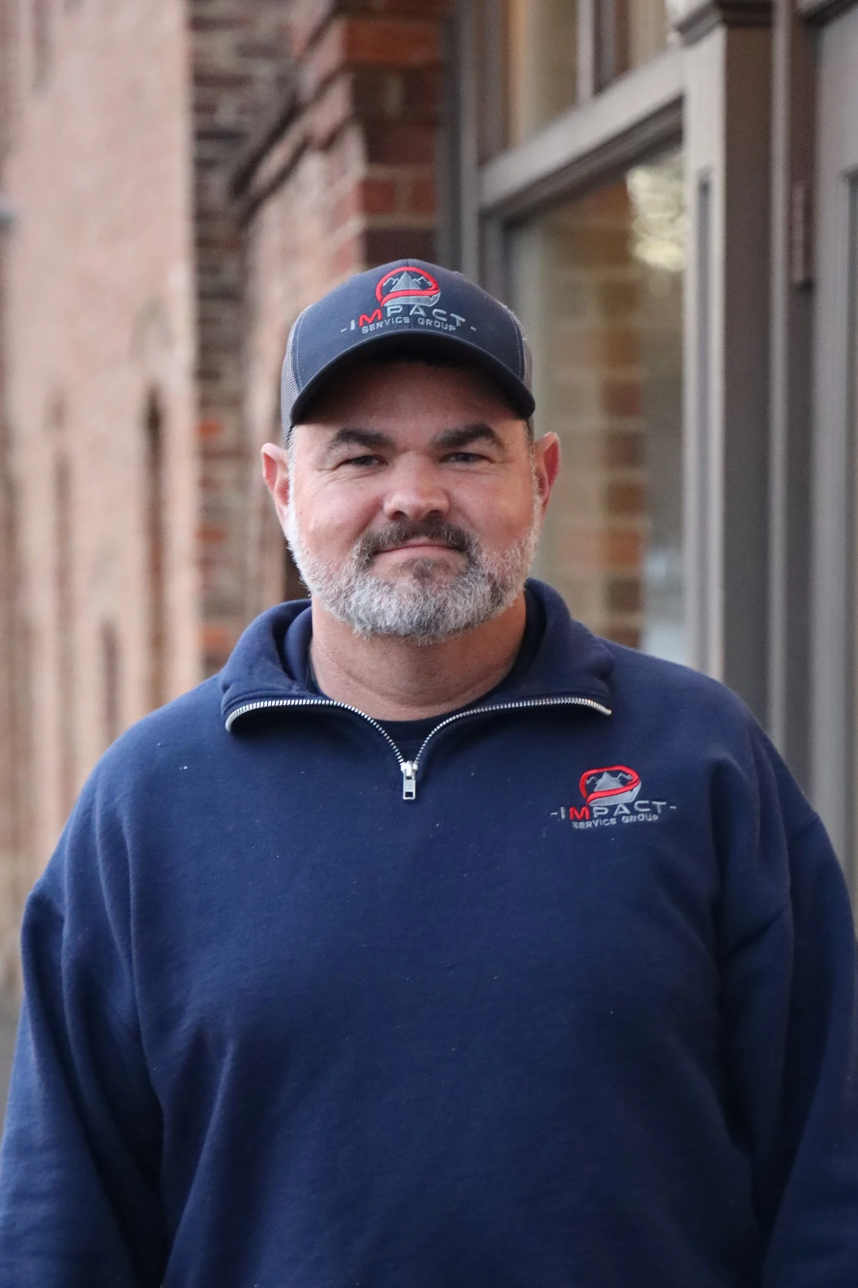 A man with a beard wearing a dark blue sweatshirt and a matching cap with the 'Impact Service Group' logo, standing outdoors in front of a brick wall and a building with large windows.
