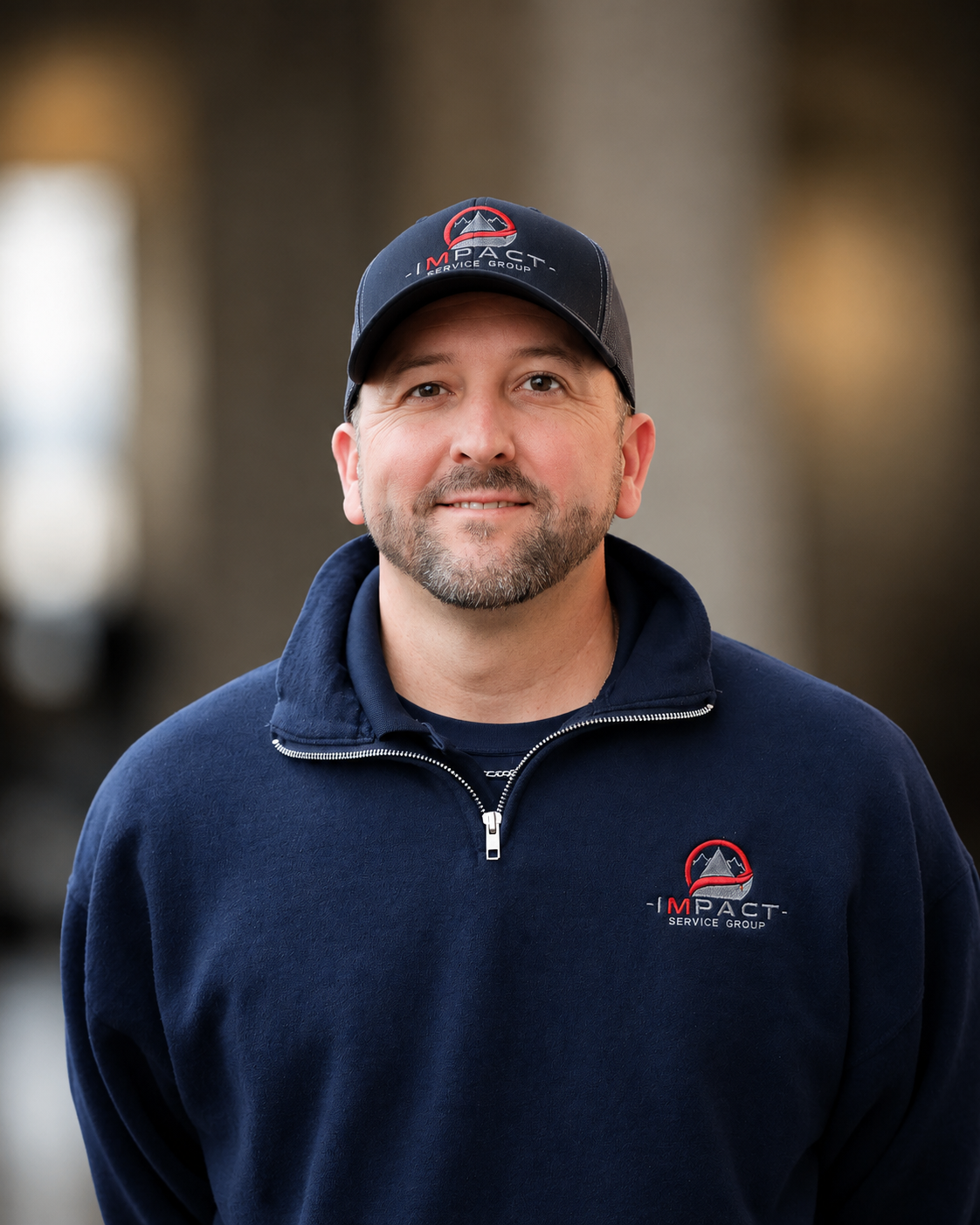 Man wearing a navy blue Impakt Service Group sweatshirt and cap, standing outdoors near a building with brick and metal siding.