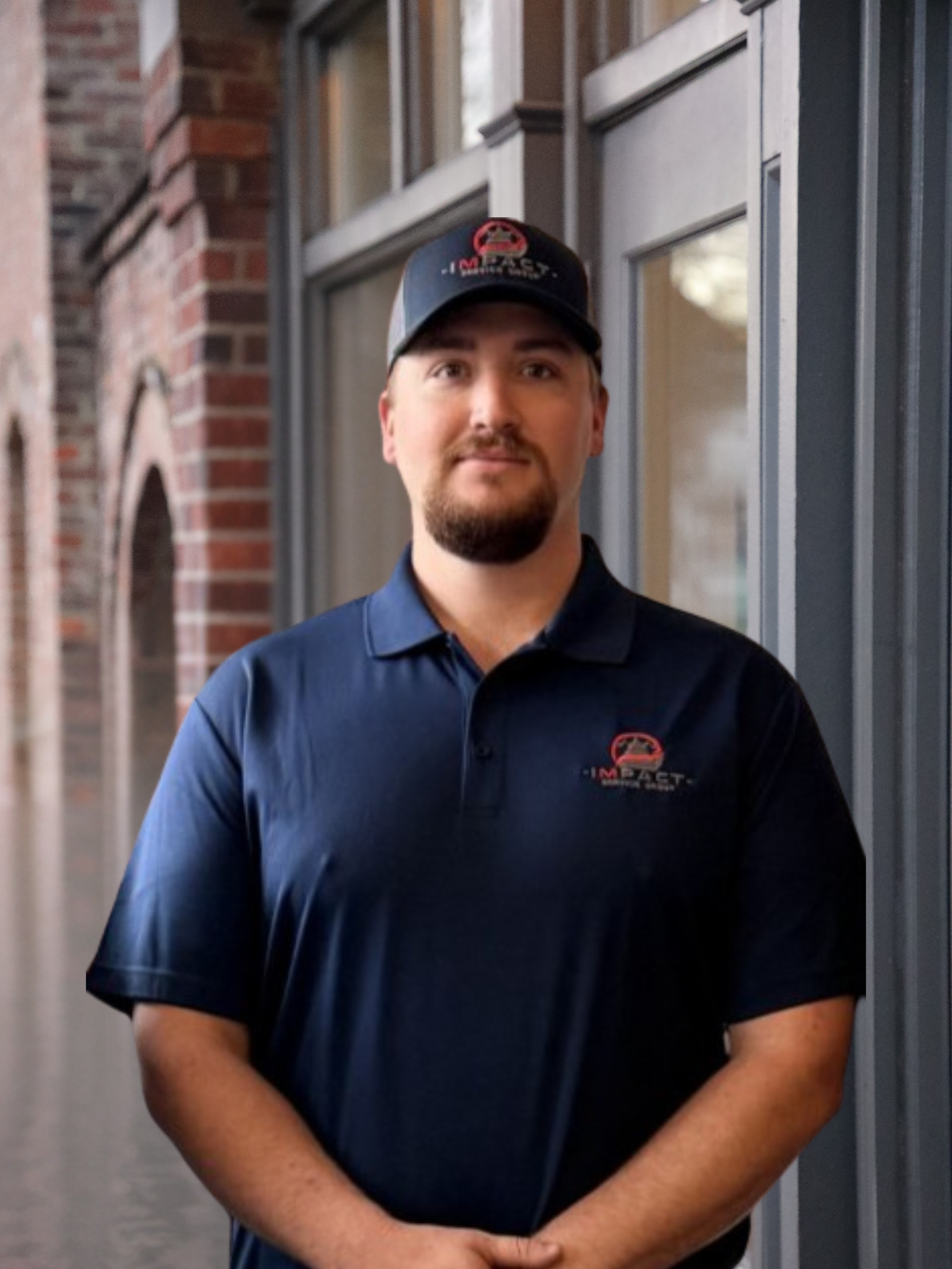 A man in a navy blue polo shirt and matching cap with a logo from Impact Safety Co. standing outside in front of a brick building with large windows.