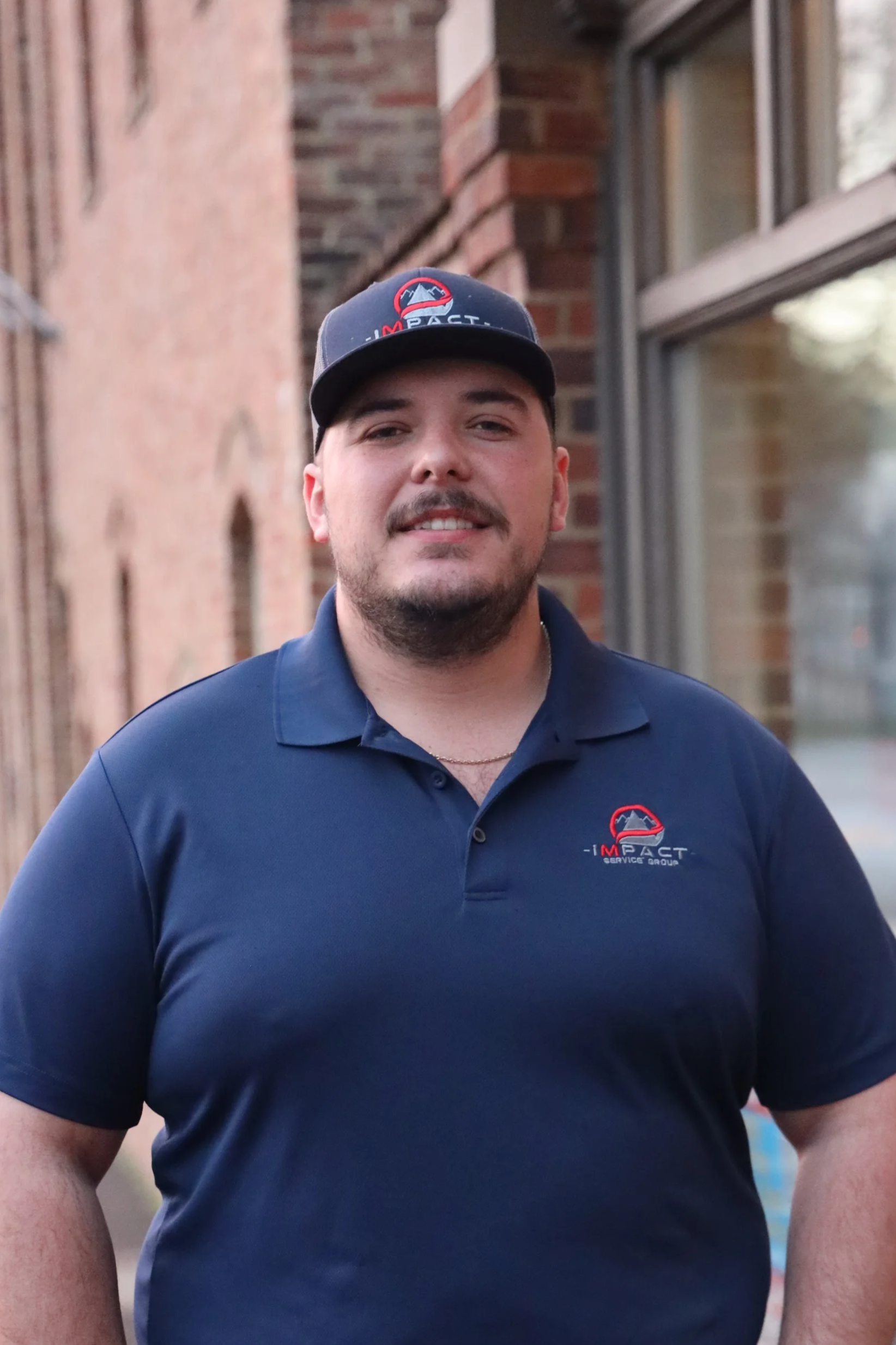 A man wearing a navy blue polo shirt and a baseball cap with a logo, standing outside near brick walls and a window.