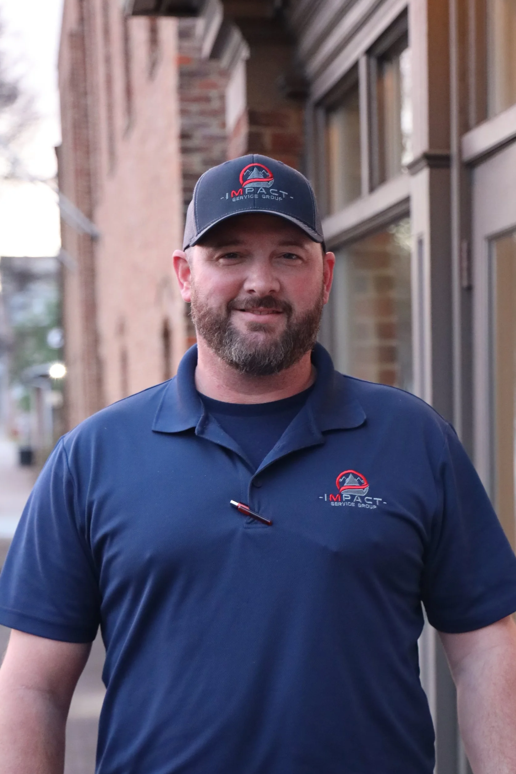 A man with a beard and short hair wearing a navy blue polo shirt and a matching cap, both with the 'Impact Service Group' logo, standing outside on a sidewalk with a brick building and windows in the background.