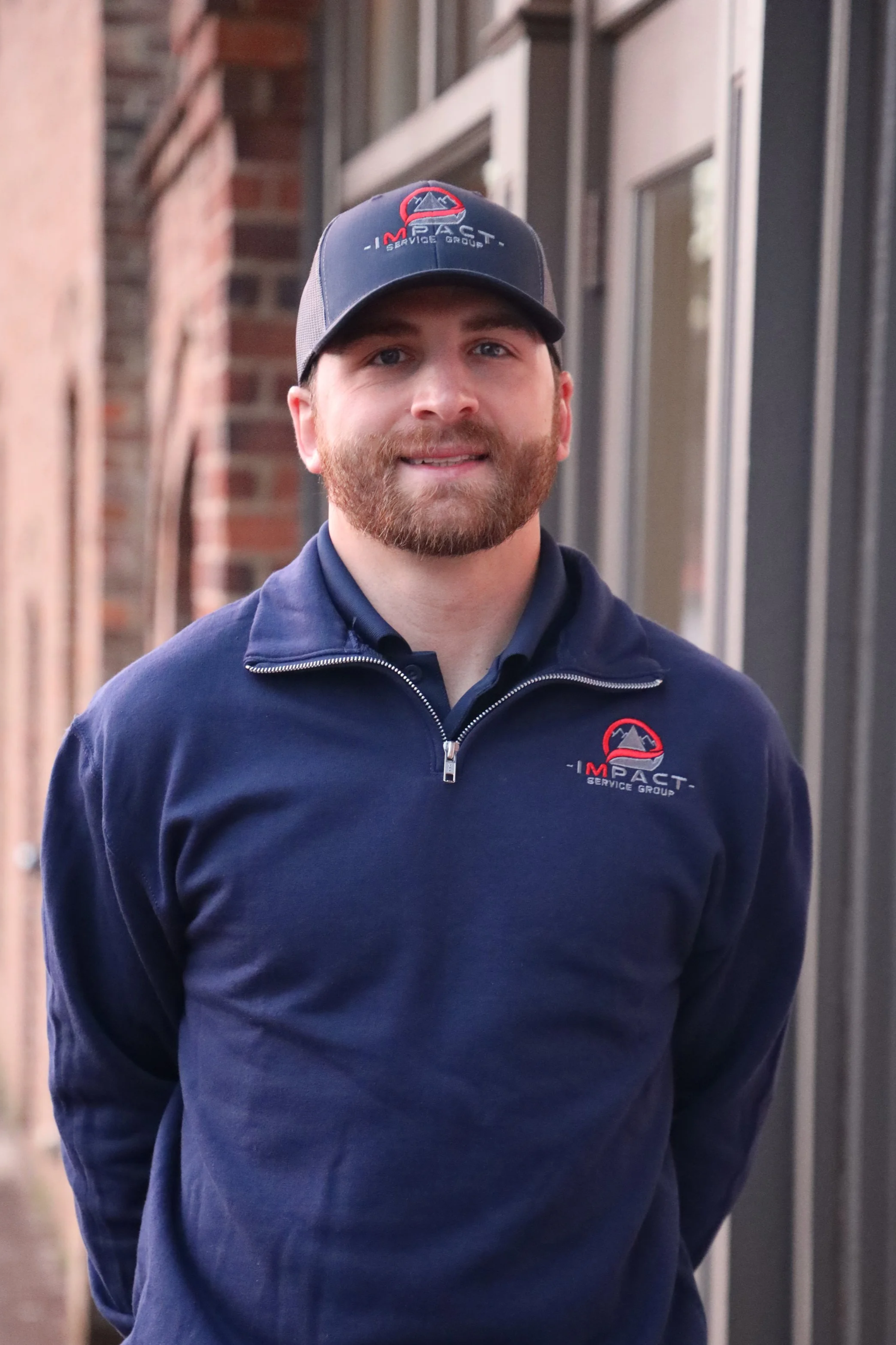 A man with a beard wearing a navy blue jacket and a cap with an 'Impact Service Group' logo, standing outdoors near a brick building.
