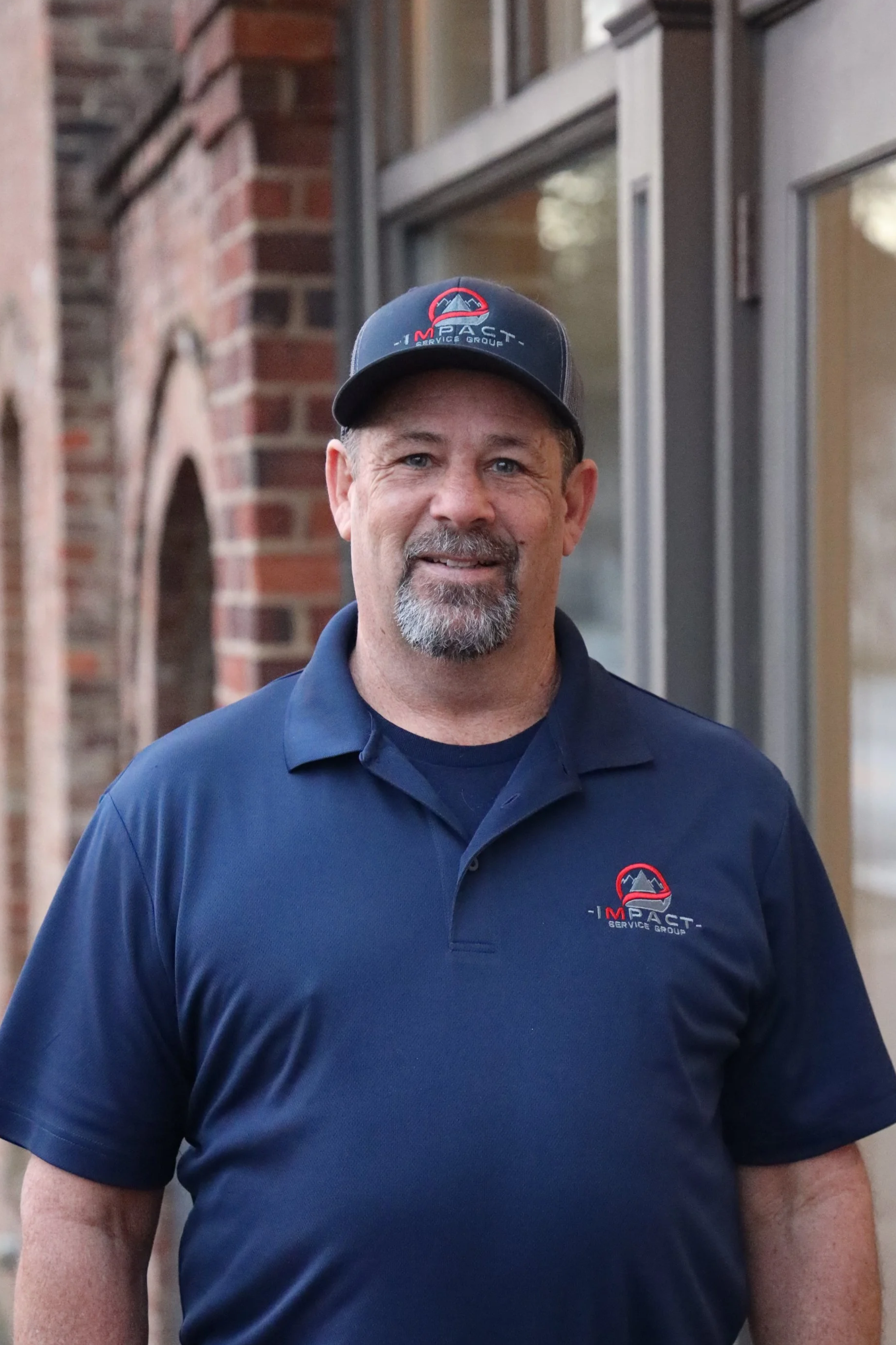 A man with a beard and mustache wearing a dark cap and navy polo shirt, both with the 'Impact Service Group' logo, standing outdoors near a brick building with large windows.