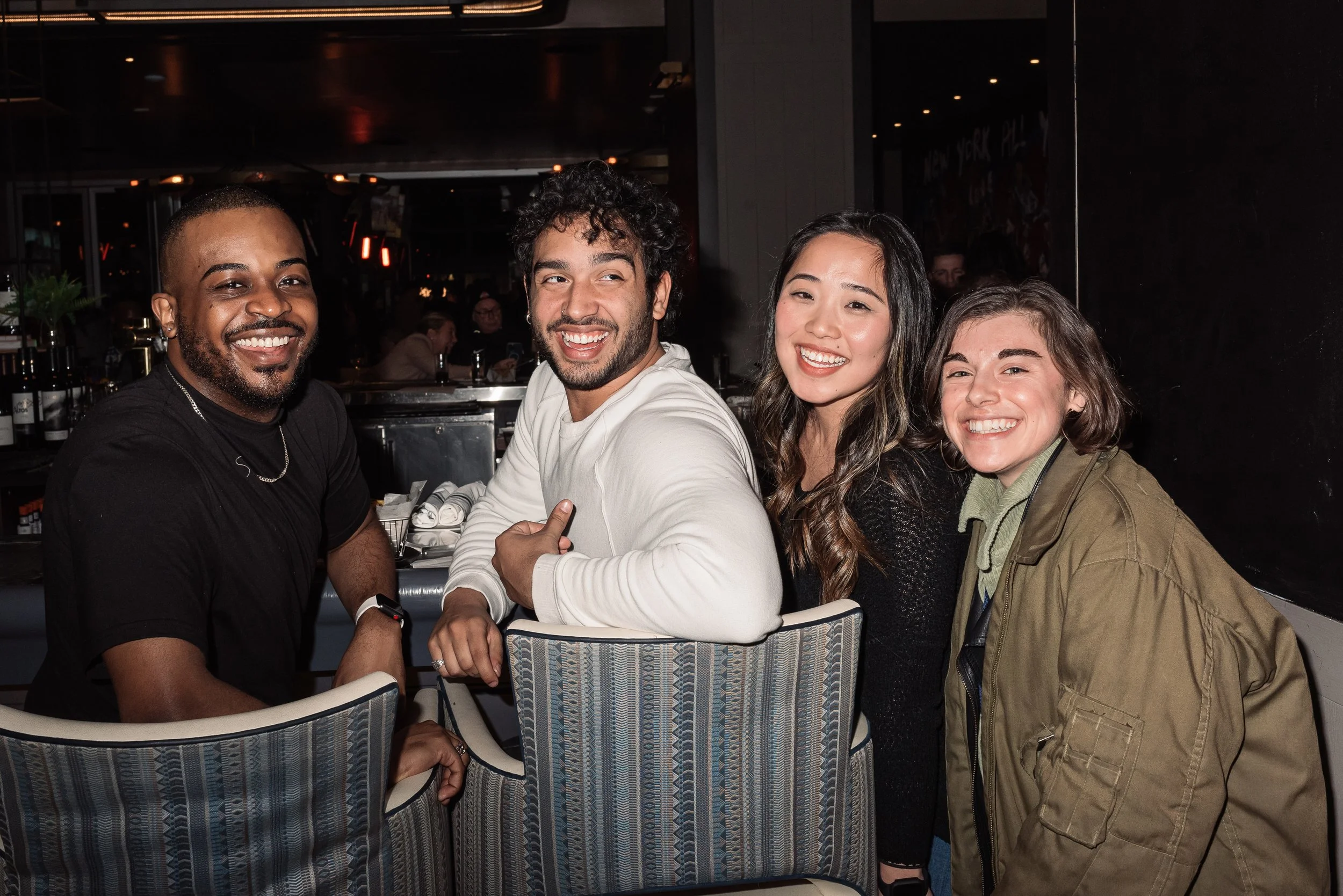 Four friends smiling at a restaurant, seated next to a dark wall, with drinks and bottles visible in the background.