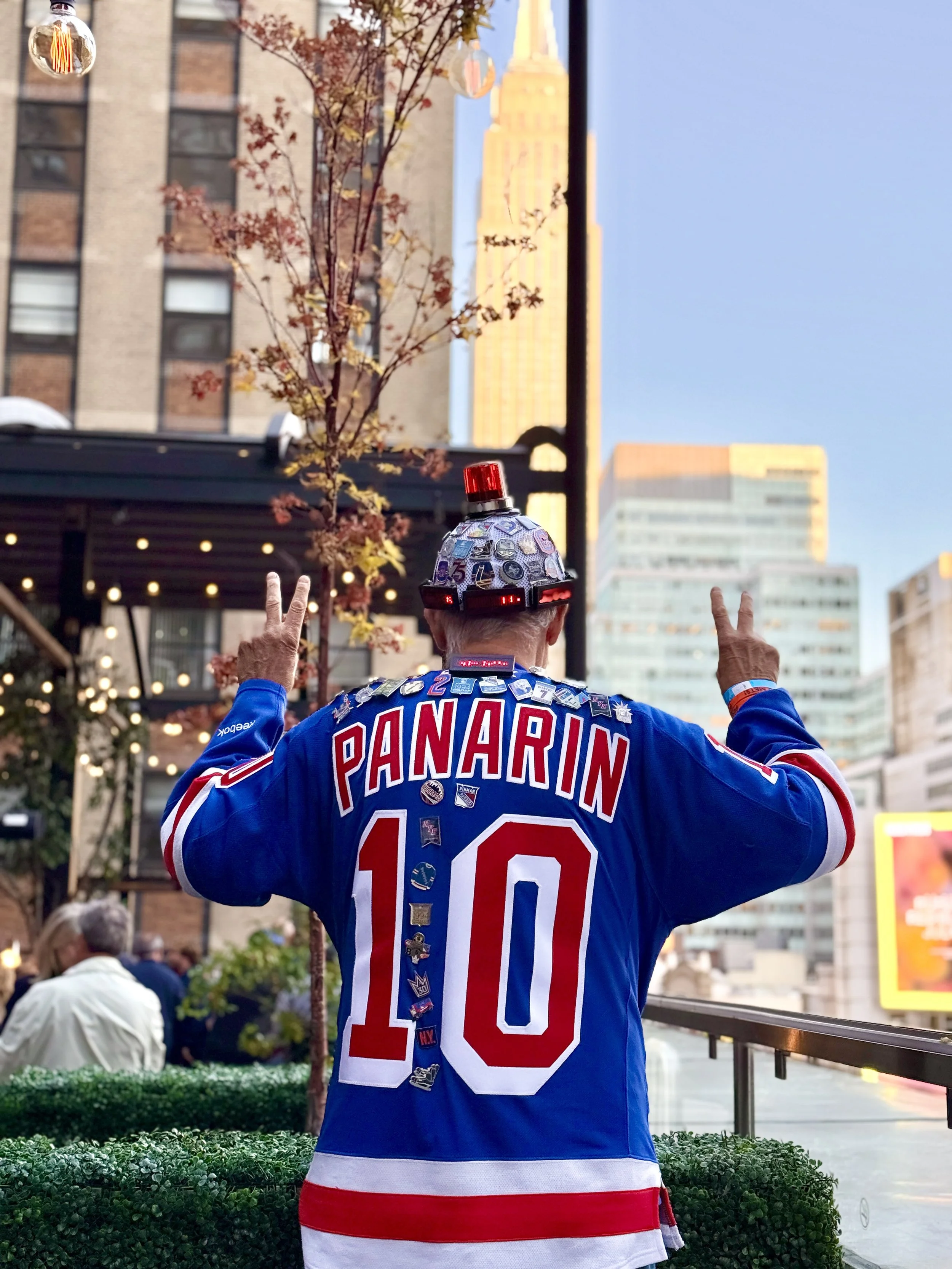 Person from behind wearing a New York Rangers hockey jersey with the name 'Panarin' and the number 10, making peace signs with both hands during sunset in an urban setting.