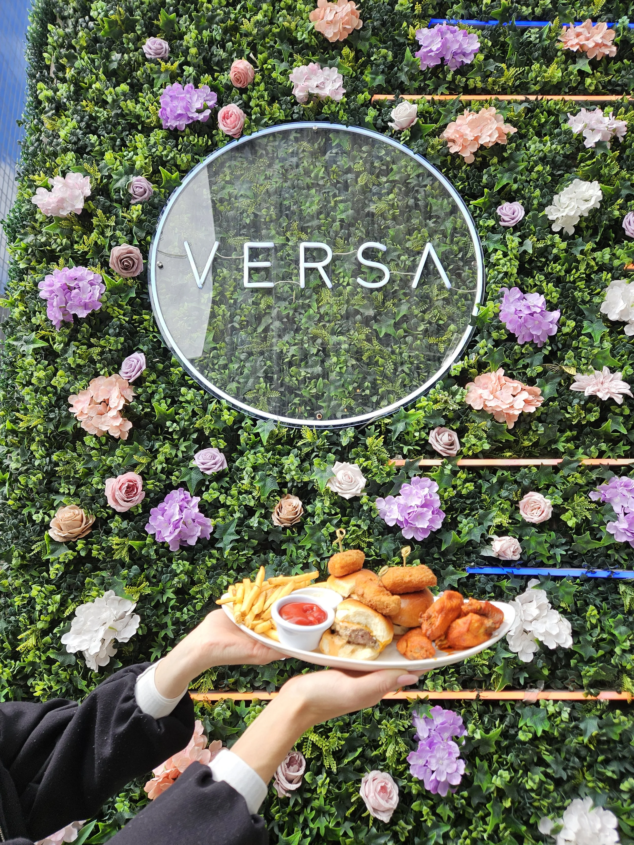 A person holding a plate of fried foods, French fries, and dipping sauces in front of a decorative wall covered with colorful artificial flowers and greenery, including roses and hydrangeas, with a neon sign that says 'VERSA'.