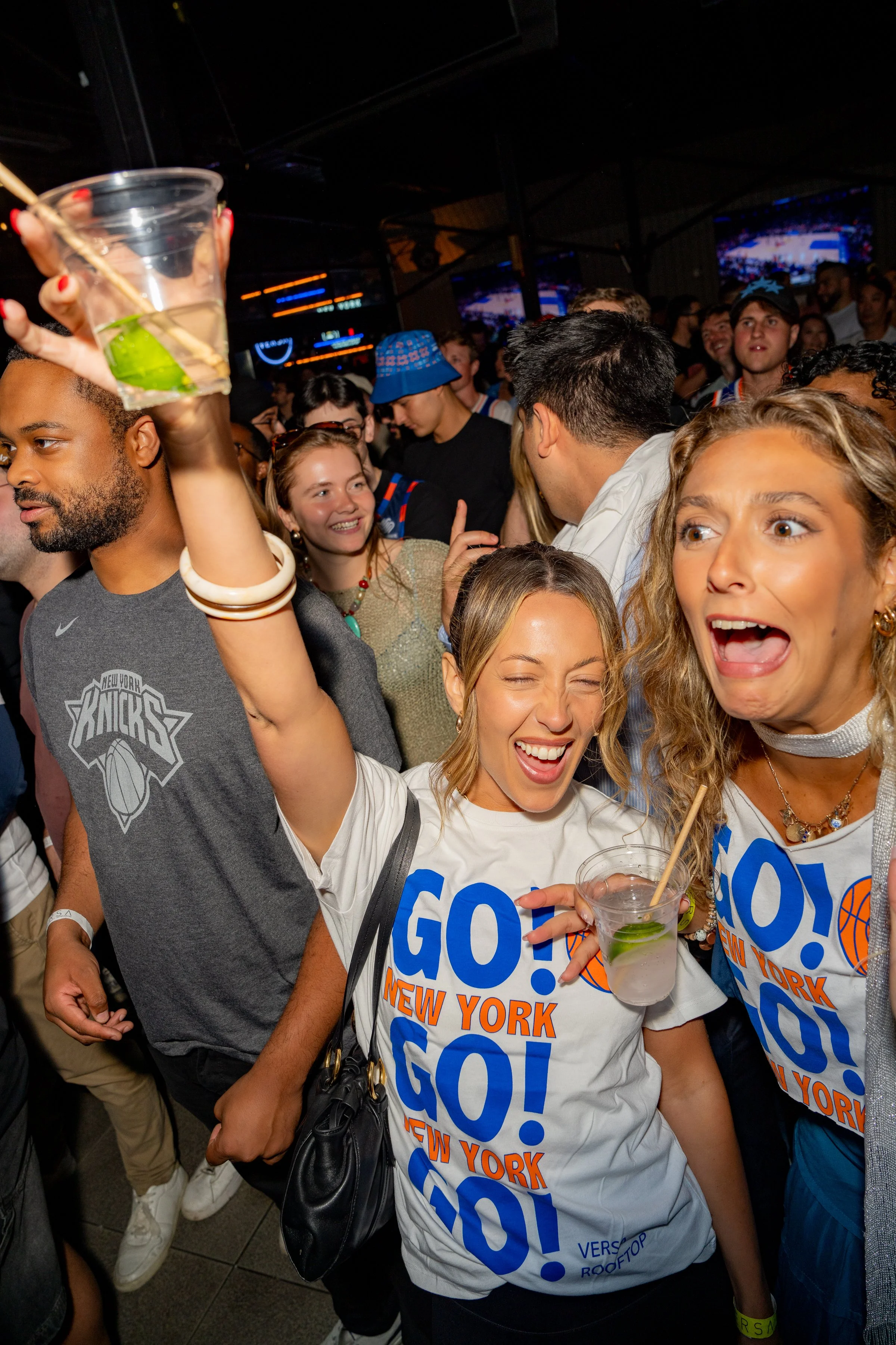 People celebrating at a New York Knicks event, wearing blue and white shirts with 'GO! NEW YORK' printed on them, holding drinks, and enjoying a lively atmosphere.