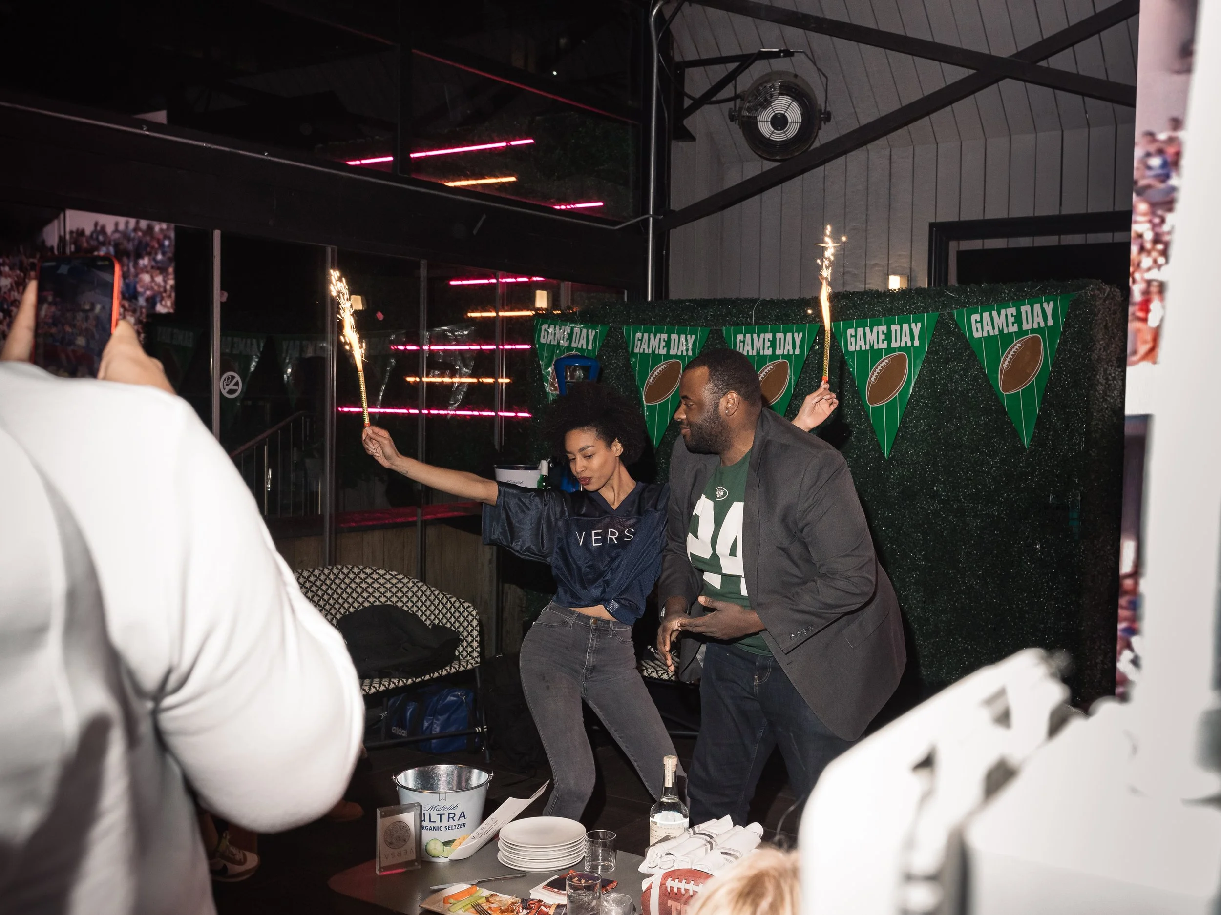 Two people celebrating at an indoor gathering with football-themed decorations and sparklers, with a sign reading 'Game Day' and football banners in the background.