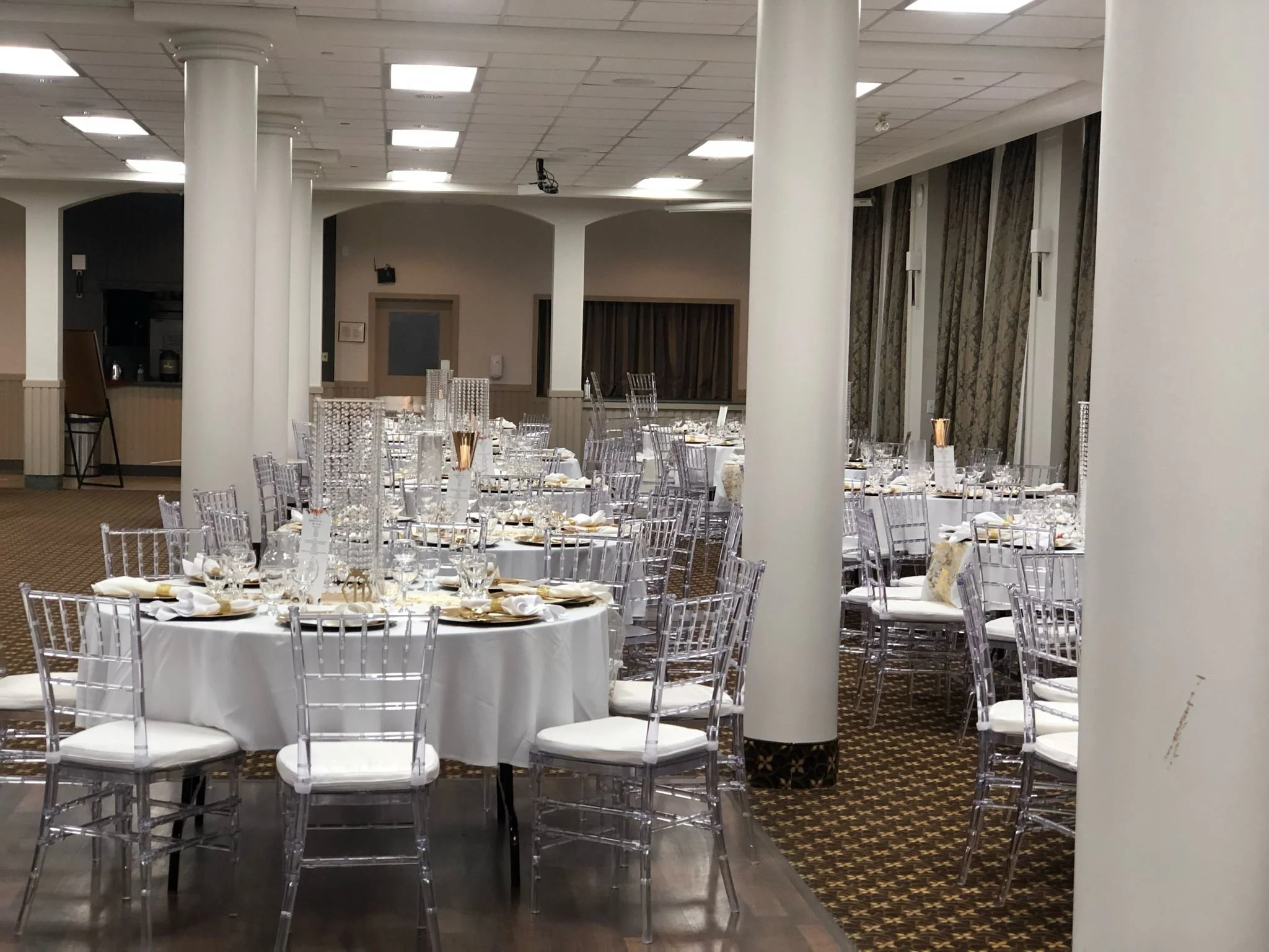 Elegant banquet hall set up with round tables covered in white tablecloths, transparent chairs with white cushions, and gold-accented tableware, ready for a formal event.