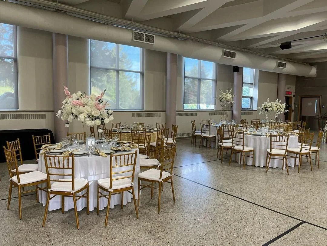 An event hall decorated with round tables draped in white cloth and adorned with large floral centerpieces. The floral arrangements feature white and pink flowers. Gold chairs with white cushions surround each table. Large windows let in natural ligh