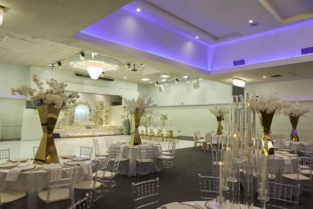 Elegant banquet hall decorated with tall floral centerpieces, round tables with white tablecloths and chairs, and blue lighting on the ceiling.