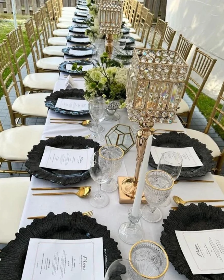 Elegant outdoors banquet table set with gold chairs, black ruffled plates, white napkins, gold flatware, crystal glasses, floral centerpieces, candles in crystal holders, and a white tablecloth.