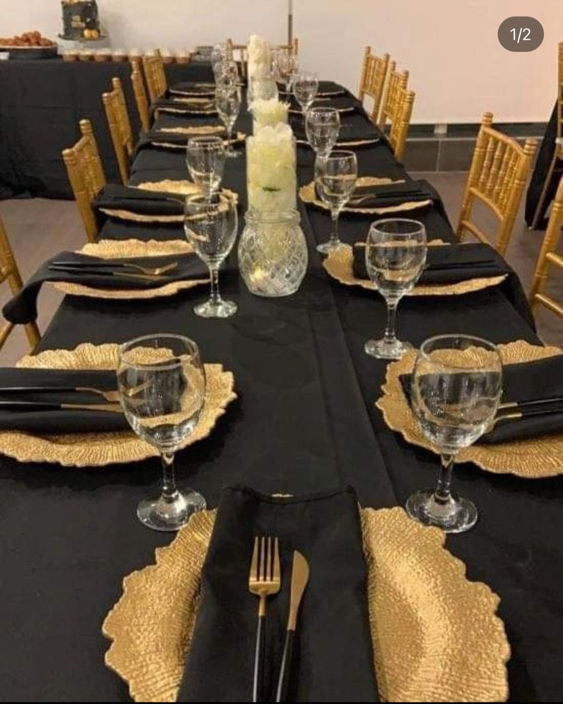 A long dining table set for a formal event with black tablecloth, gold placemats, black napkins, wine glasses, and gold-colored chairs.