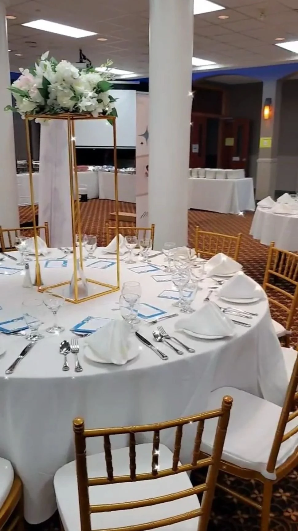 A round banquet table set with white tablecloth, folded napkins, glassware, and silverware, decorated with a tall centerpiece of white flowers in a gold geometric stand, in an event hall with other decorated tables and a large screen in the backgroun