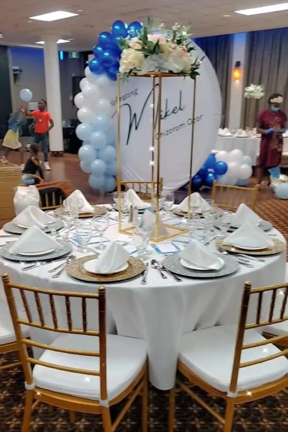 Elegant banquet table decorated for a celebration, with a centerpiece that reads 'Wikel' and balloons in blue and white, surrounded by people in a large party hall.