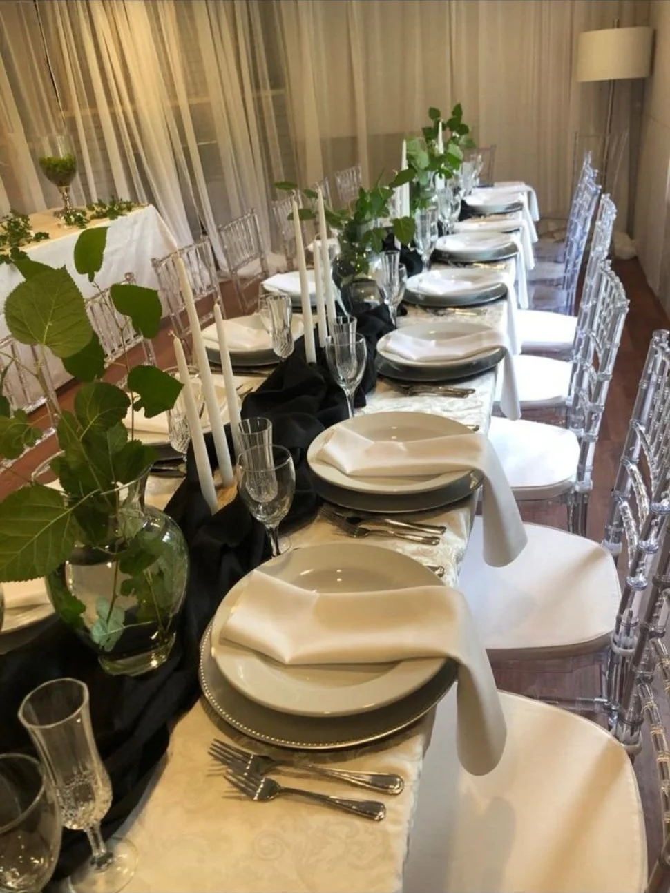 Elegant dining table set with white plates, folded napkins, silverware, wine glasses, and greenery centerpiece, with clear chairs and beige curtains in the background.