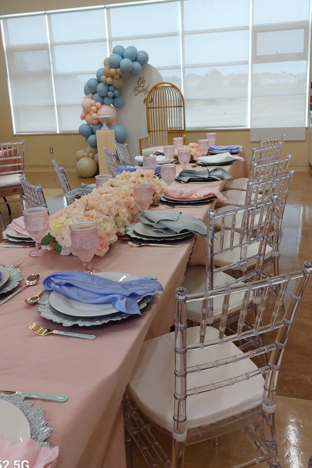 Elegant dining table decorated with pastel pink tablecloth, floral centerpiece, pink glasses, and neatly arranged plates, silverware, and napkins in front of a window with balloons and a decorative backdrop.