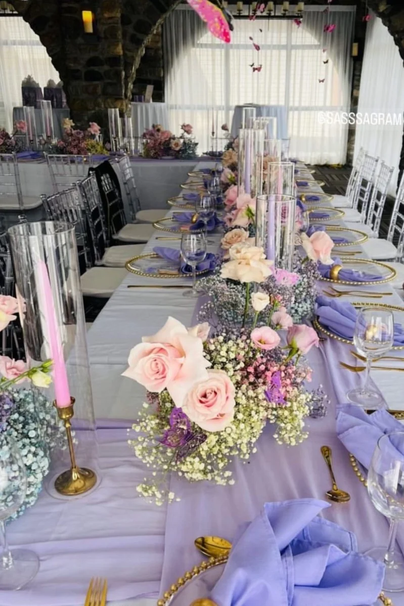 Elegant wedding reception table decorated with pastel pink and purple flowers, gold cutlery, and purple napkins, with tall glass candle holders and butterfly accents.