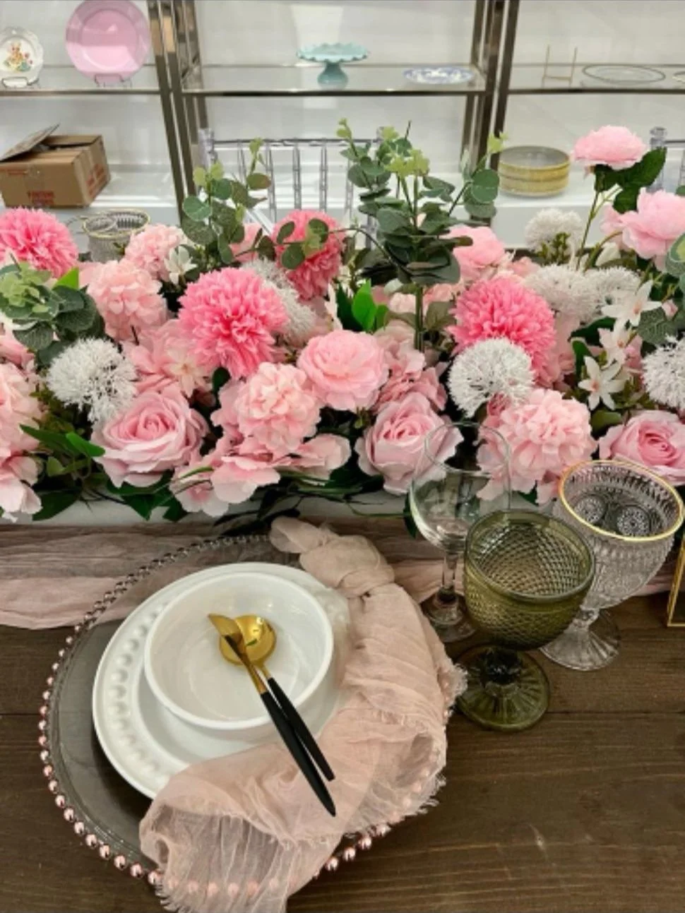 Table setting with pink and white flowers, including roses and dahlias, on a wooden table. There are various glasses, a gold and black spoon on a white plate, a pink ruffled napkin, and decorative dishes in the background.