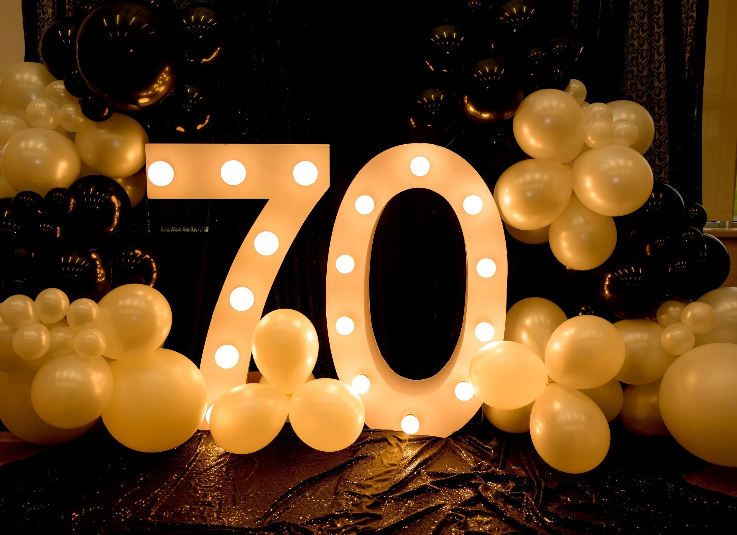 Decorative display celebrating a 70th birthday with large illuminated number '70', surrounded by black and gold balloons, on a shiny black fabric background.