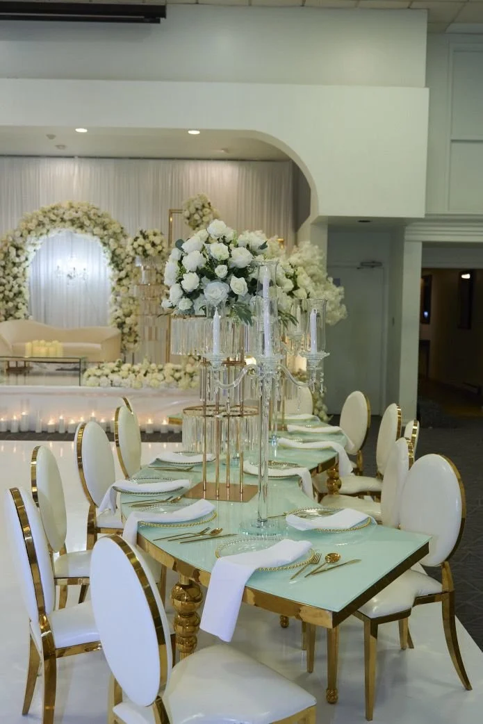 Elegant wedding reception table with white and gold chairs, floral centerpieces, and a decorated stage with white flowers in the background.