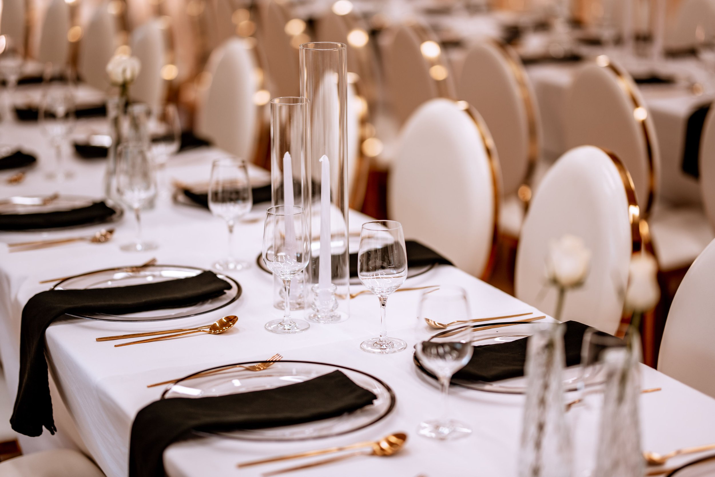 Elegant banquet table setup with white tablecloth, black napkins, gold cutlery, and clear glassware, decorated with tall glass candle holders.