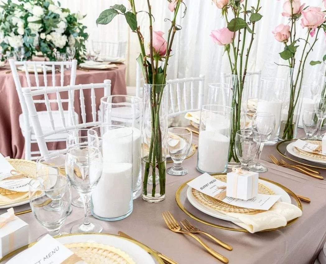 Elegant table decoration for a wedding or event with pink roses, white candles, gold cutlery, and place settings on a soft pink tablecloth.