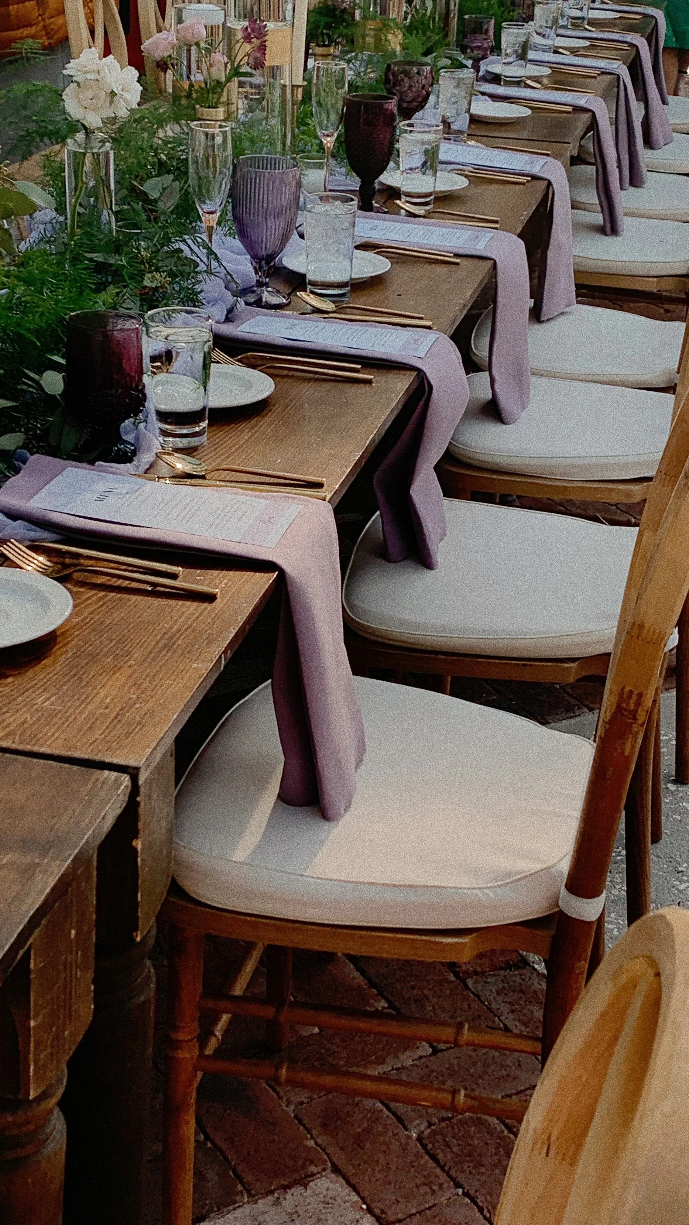 A long outdoor dining table set for a meal with purple and white glassware, gold utensils, white plates, and pastel pink and purple napkins. The table is decorated with flowers and greenery, and the chairs have white cushions. The table is placed on a brick surface.