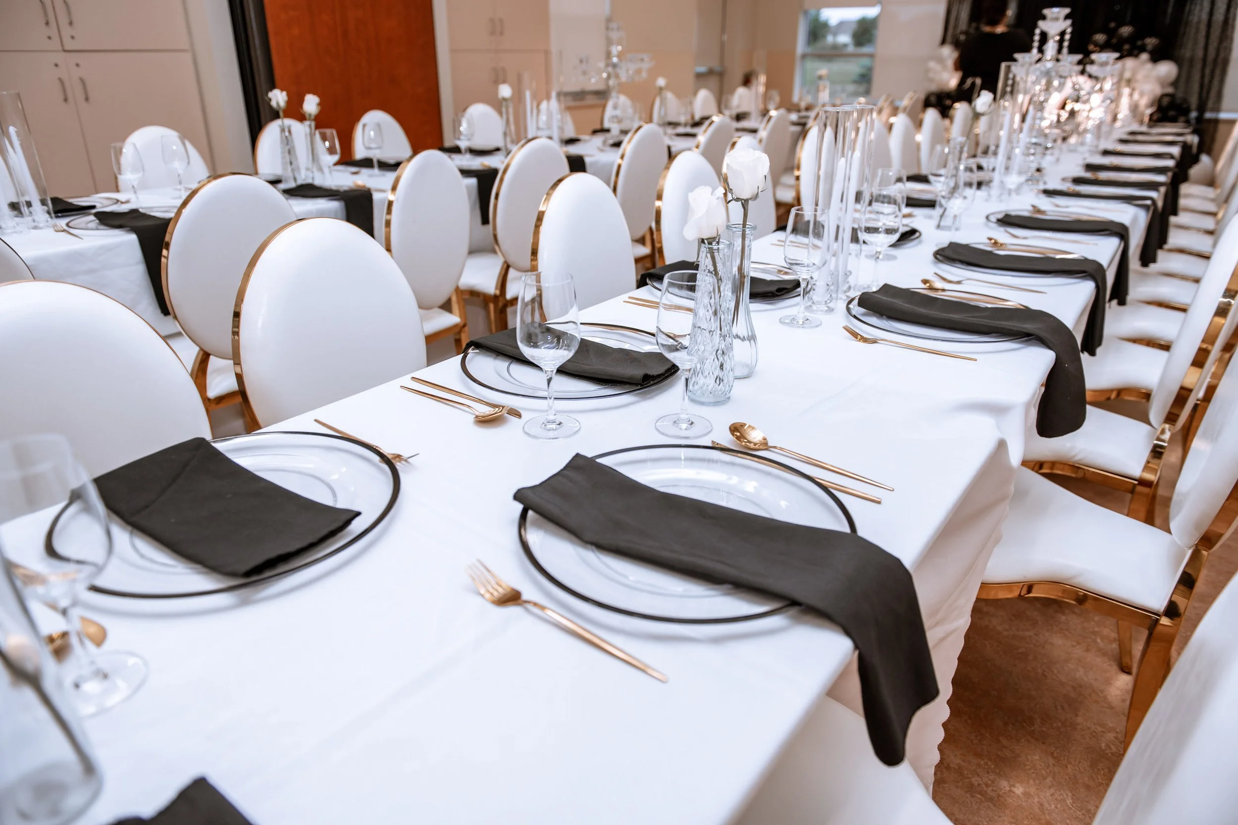 Elegant banquet table setup with white tablecloths, black napkins, gold flatware, clear glasses, and white roses in glass vases, arranged for a formal event.